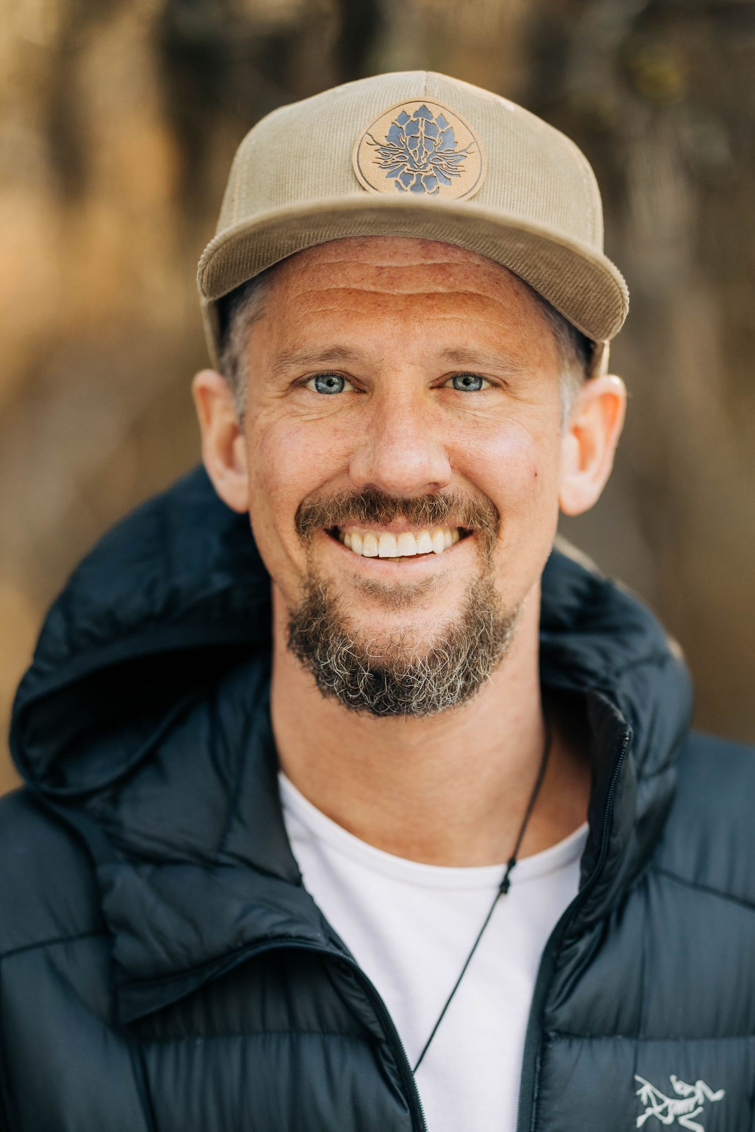 Smiling bearded man in cap and black jacket outdoors.