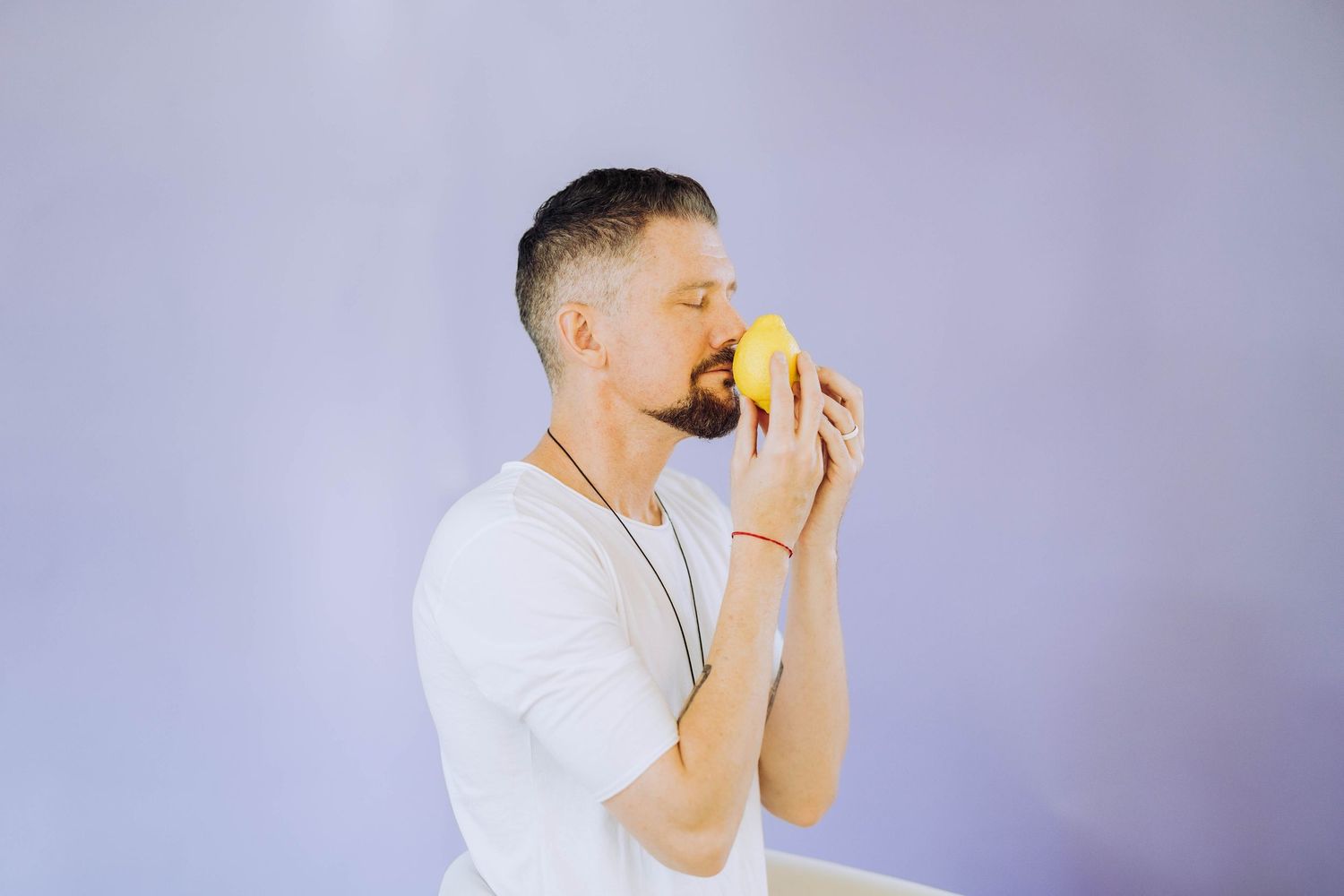 Bearded man in white t-shirt smelling a fresh yellow lemon on a light purple background.