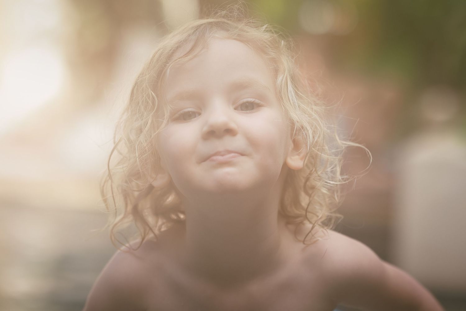 Close-up portrait of a curly blonde toddler smiling softly in warm glowing sunlight outdoors.