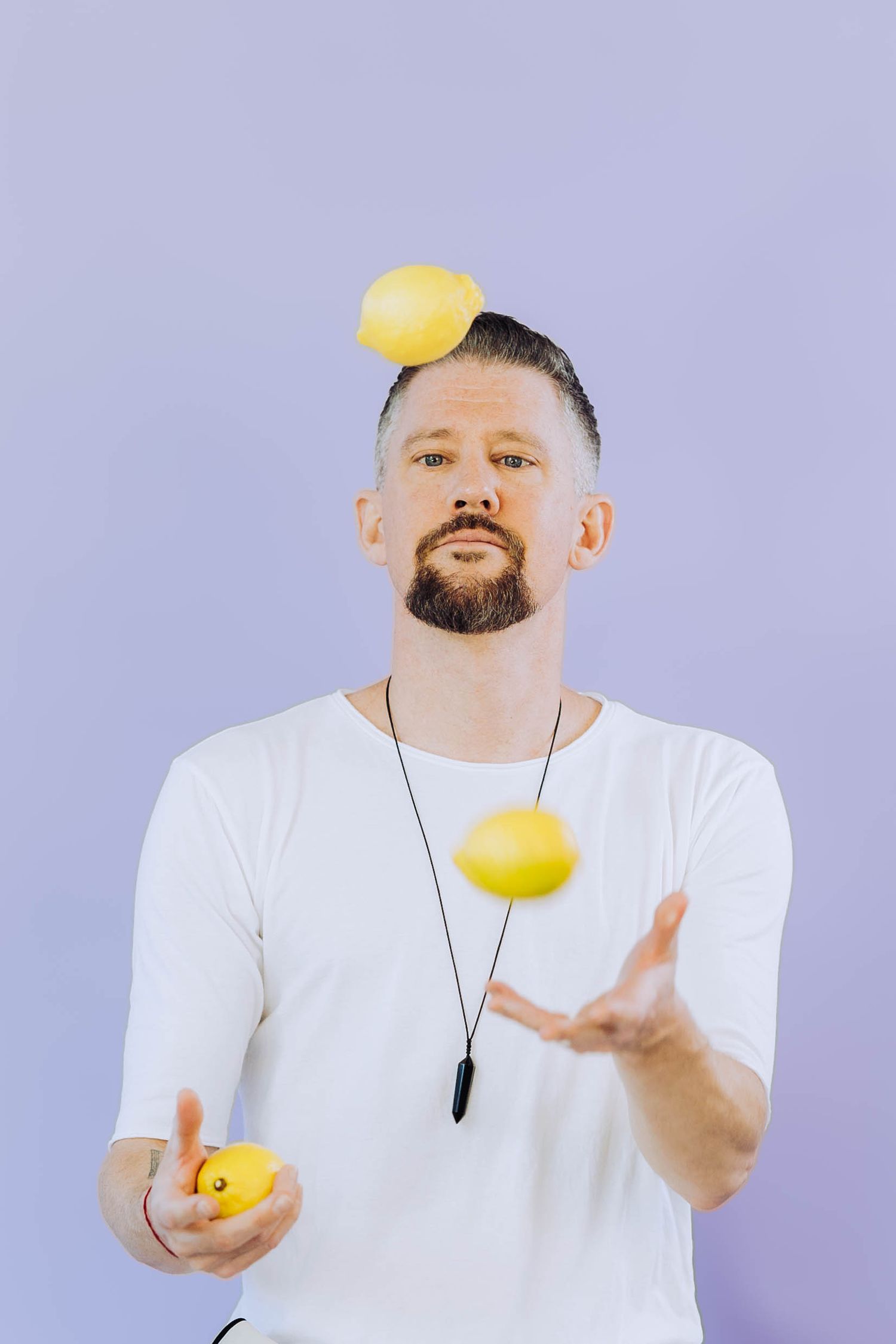 Bearded man in white t-shirt juggling yellow lemons with one balanced on his head.