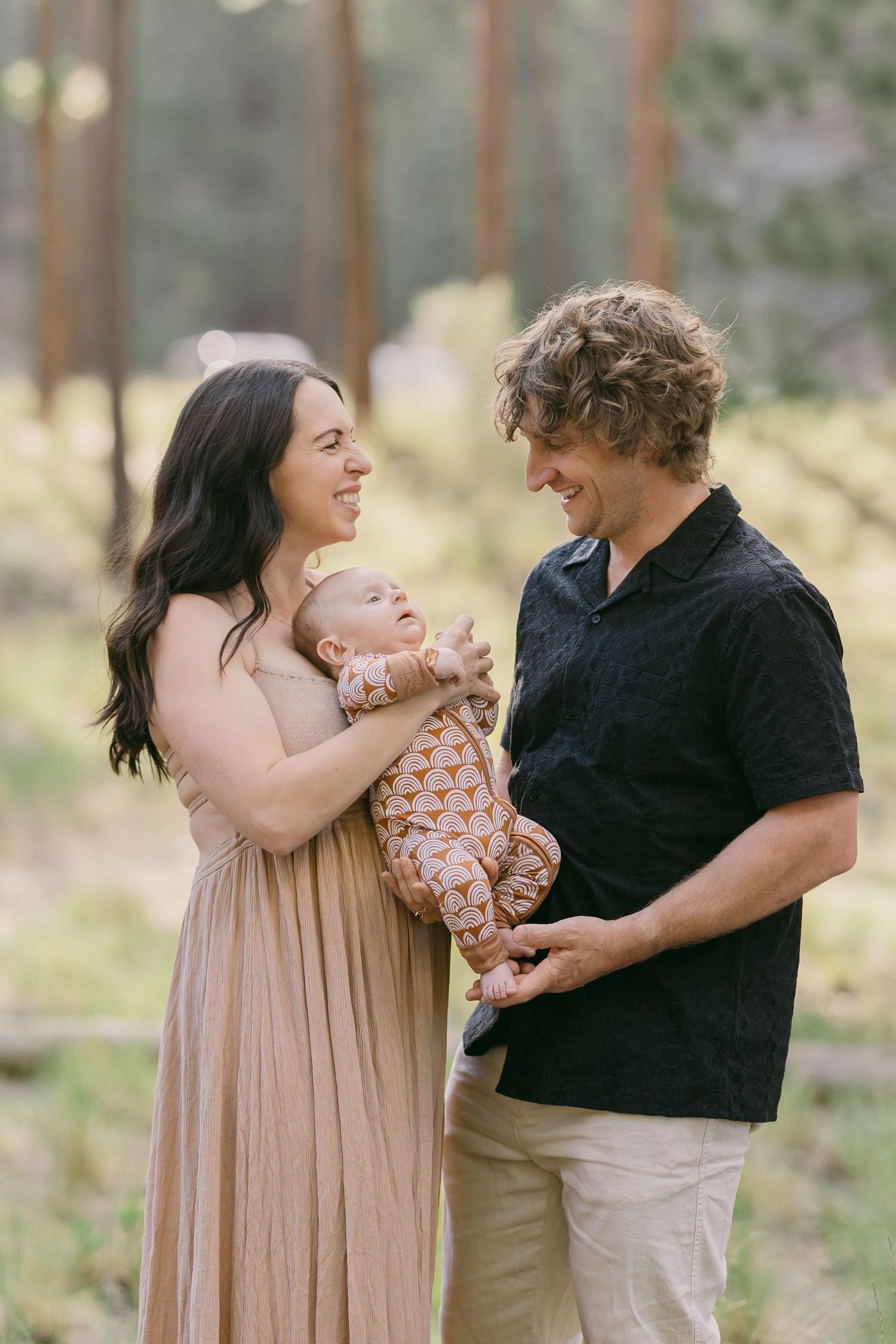 Happy young family with curly-haired man and dark-haired woman in beige dress holding newborn baby in forest.