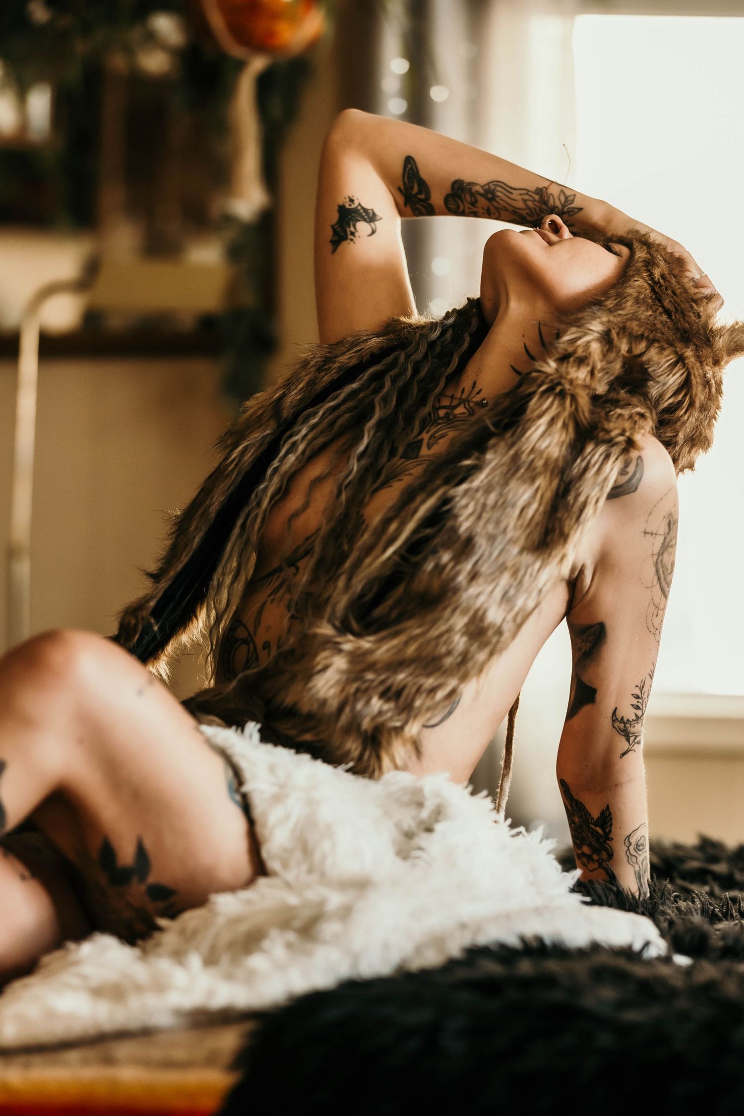 Tattooed woman posing on a fur rug wearing an open fur vest in a bohemian indoor setting.