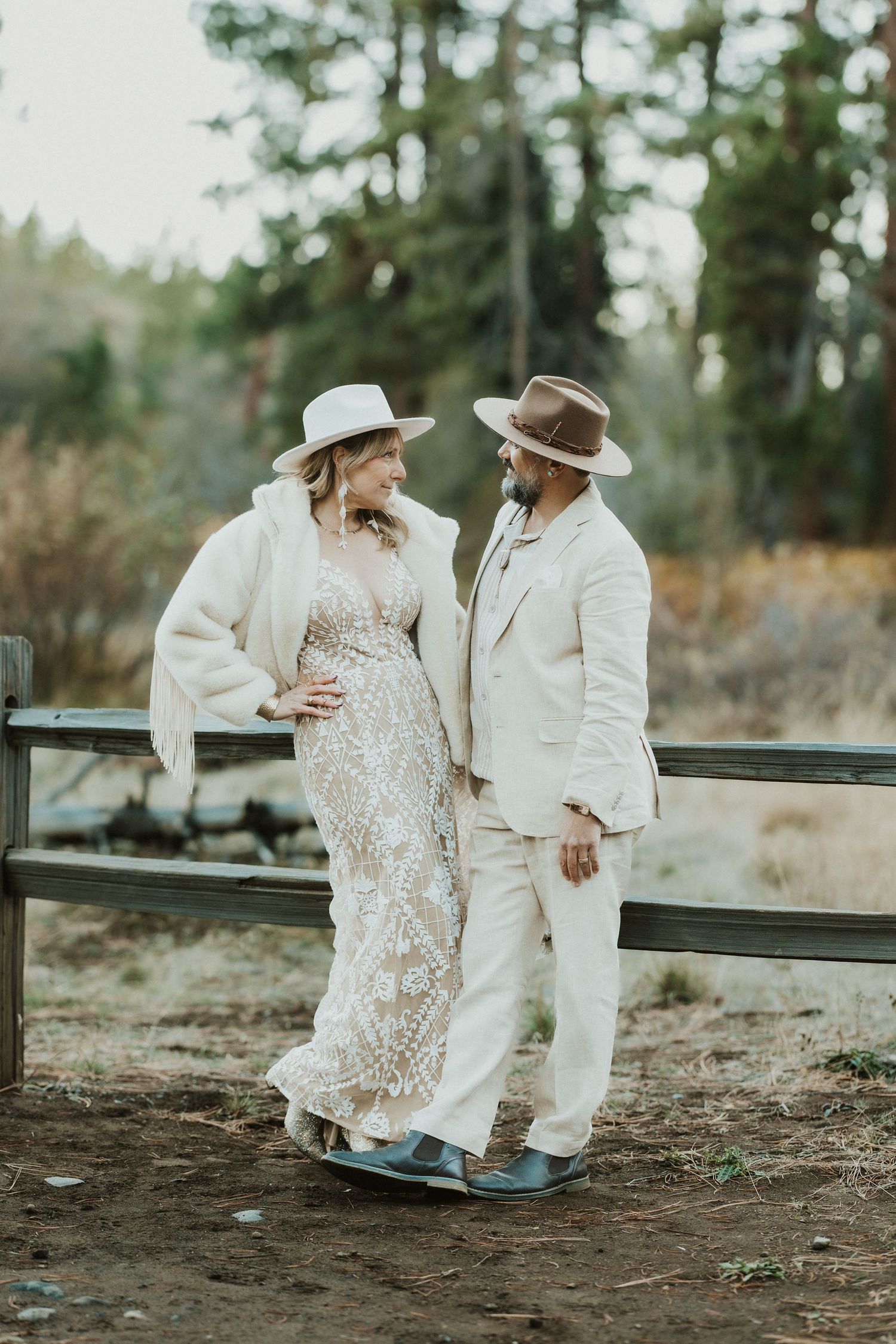 Bride in white fringe jacket and groom in cream suit lean on fence in wooded setting.