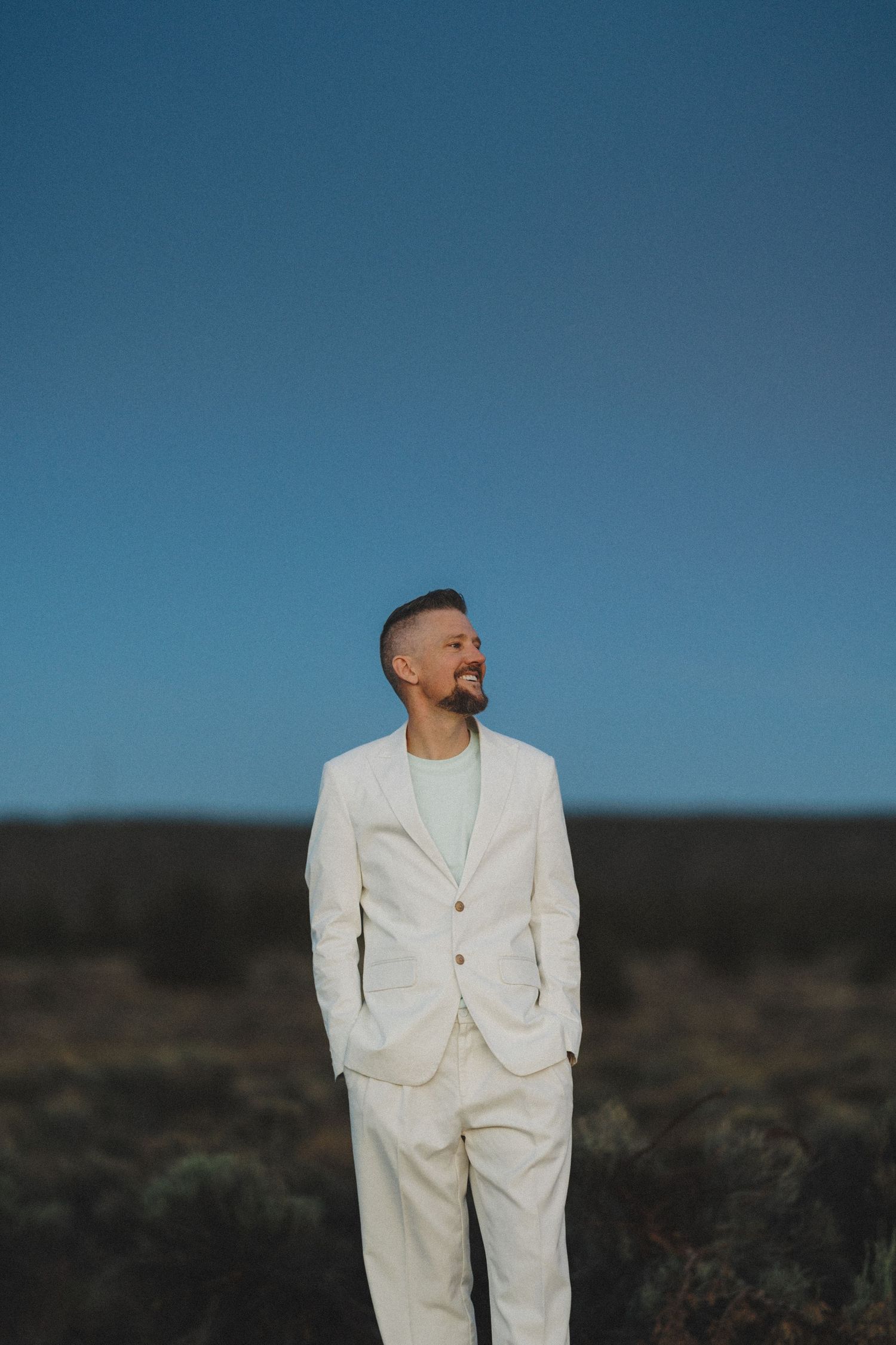 Smiling man in white suit standing in desert landscape under vivid blue sky.