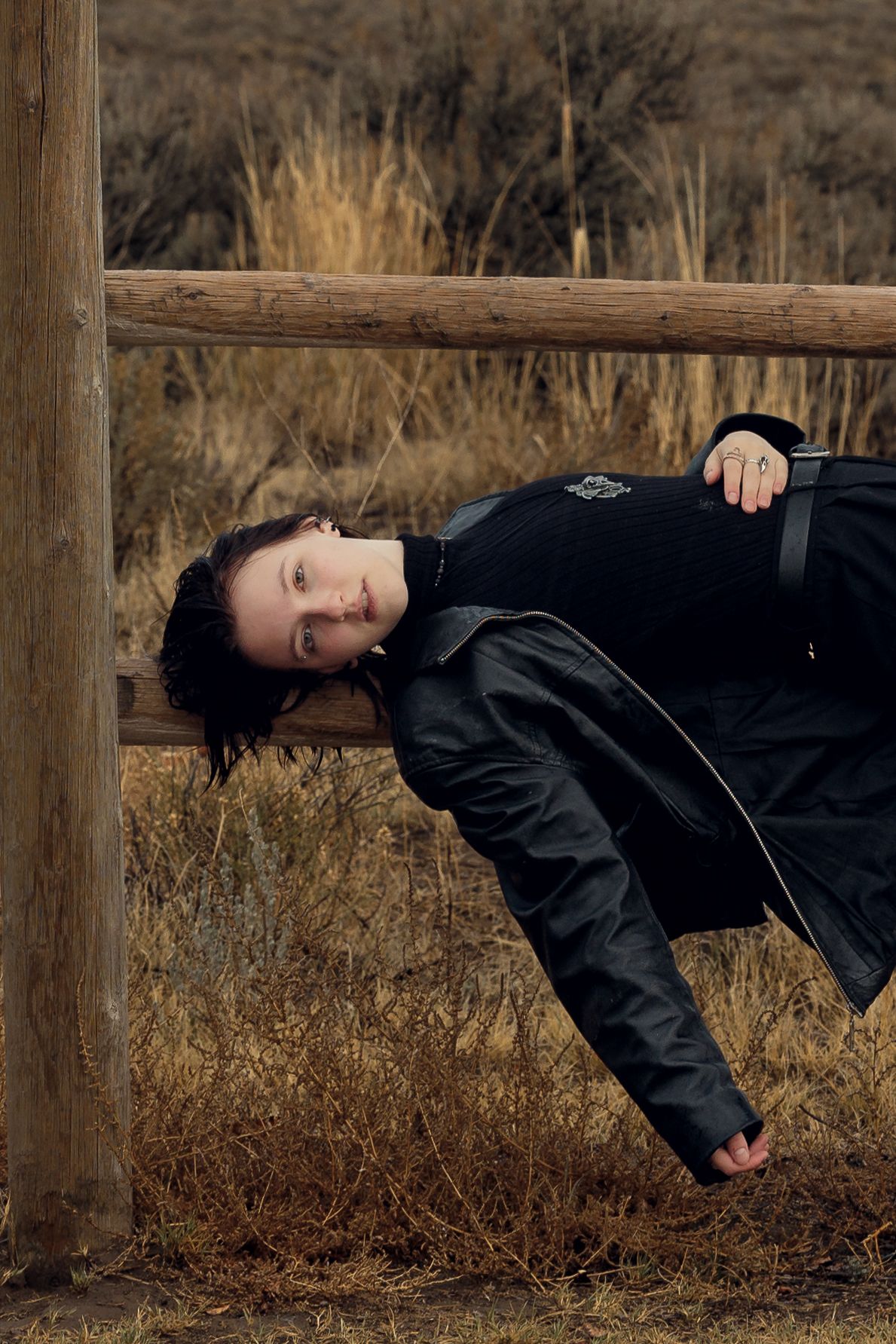 A dark-haired person in a black coat leans dramatically over a rustic wooden fence rail in a dry rural landscape.