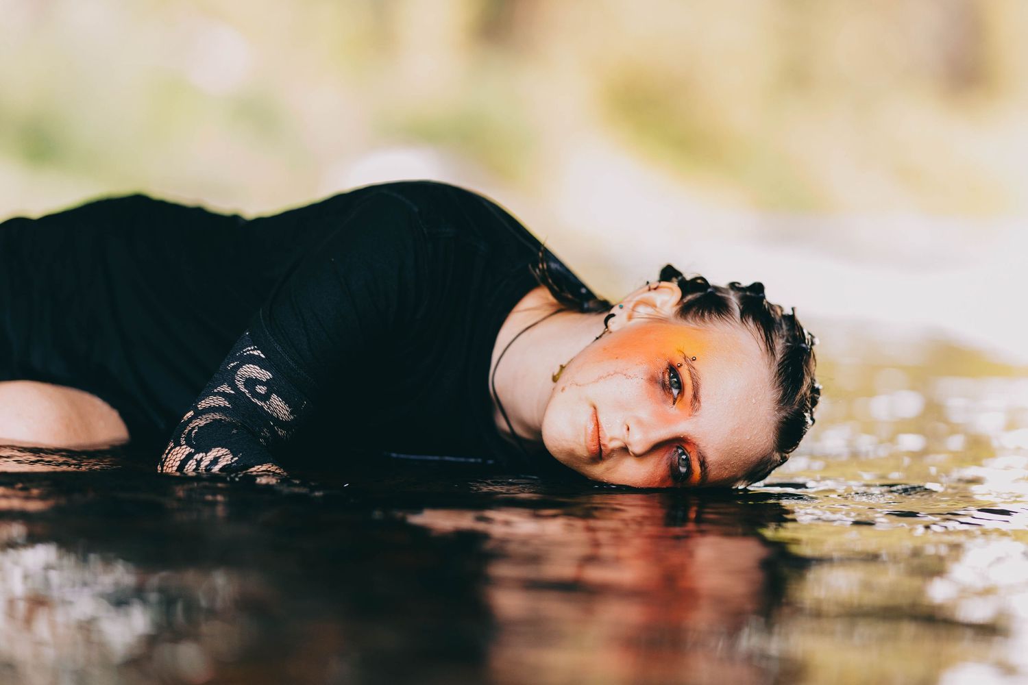 Woman with orange eye makeup lies sideways in calm water wearing black outfit.