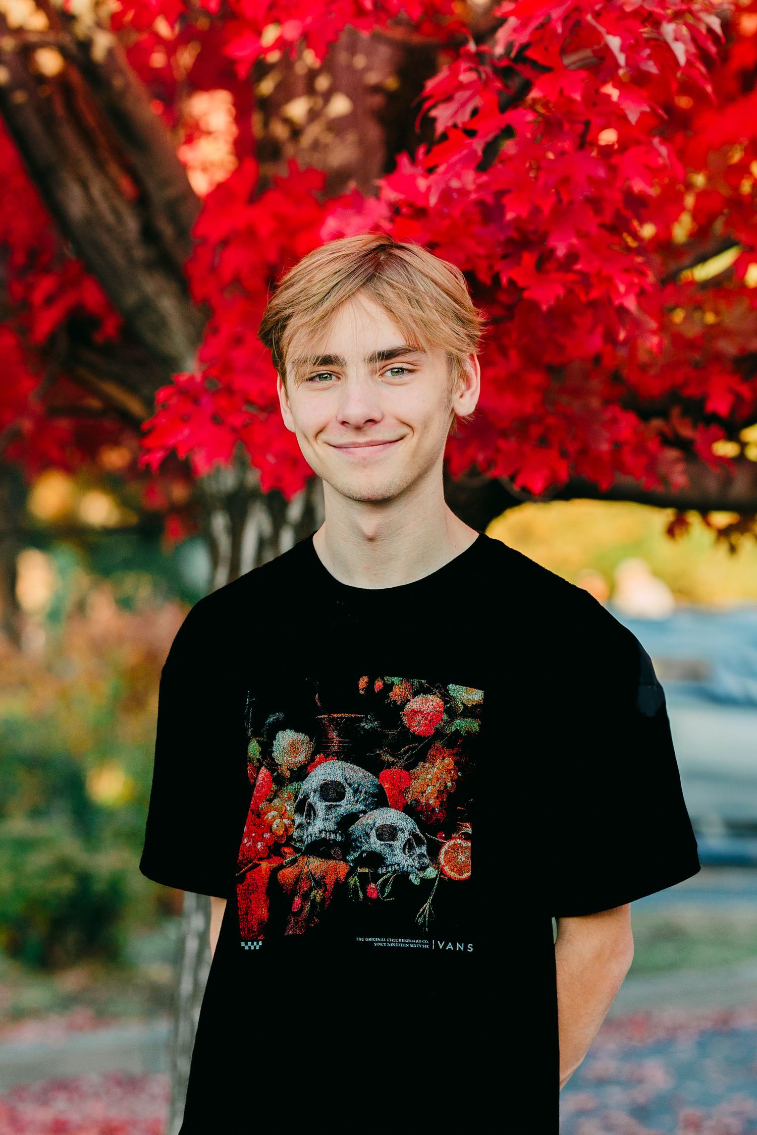 Smiling teenage boy in black Vans skull graphic tee under red autumn tree.