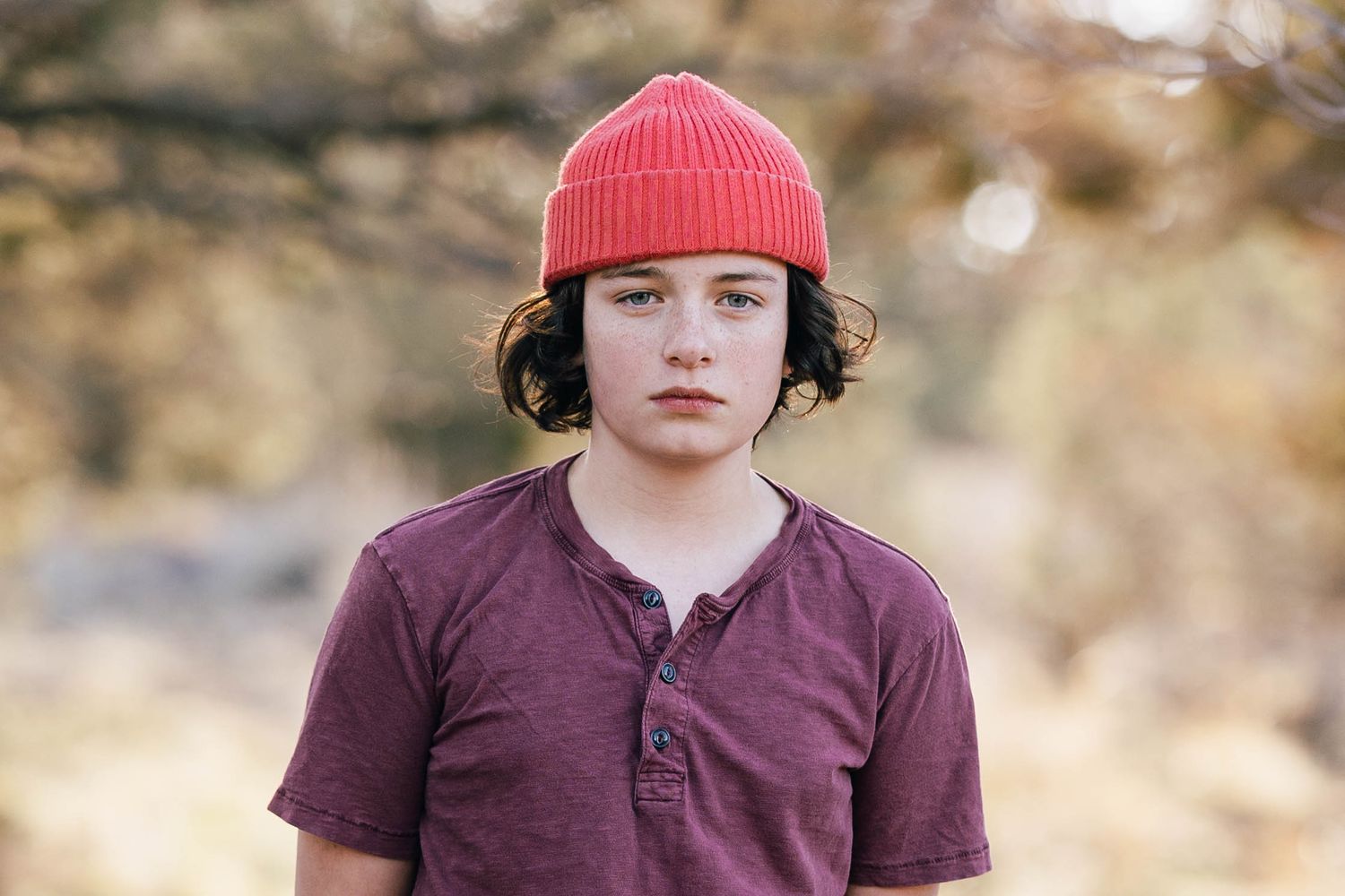 Young person in a red beanie and purple shirt standing outdoors with a serious expression.