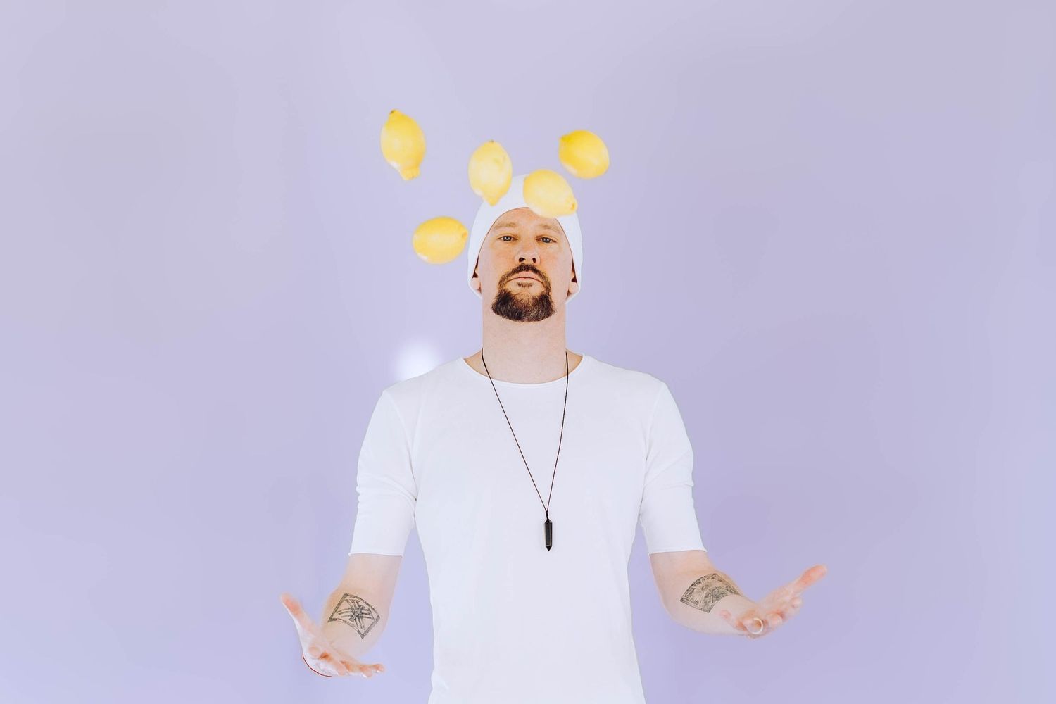 Tattooed man in white t-shirt tossing lemons in the air against a soft purple background.