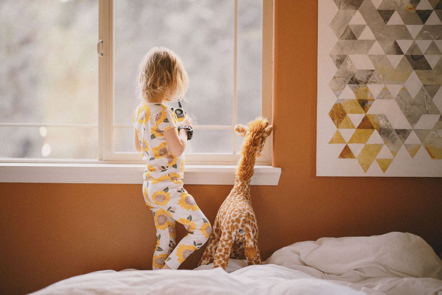 Toddler in pajamas standing on bed looking out window with giraffe toy.