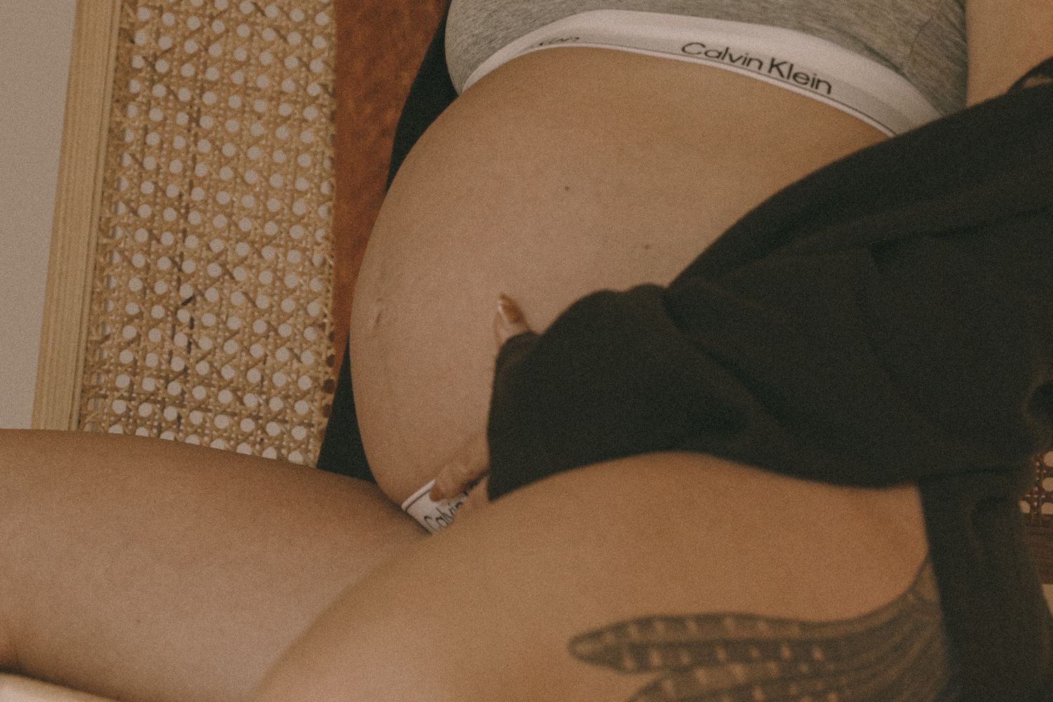 Close-up of pregnant belly in Calvin Klein underwear with tattooed thigh in casual maternity shot.