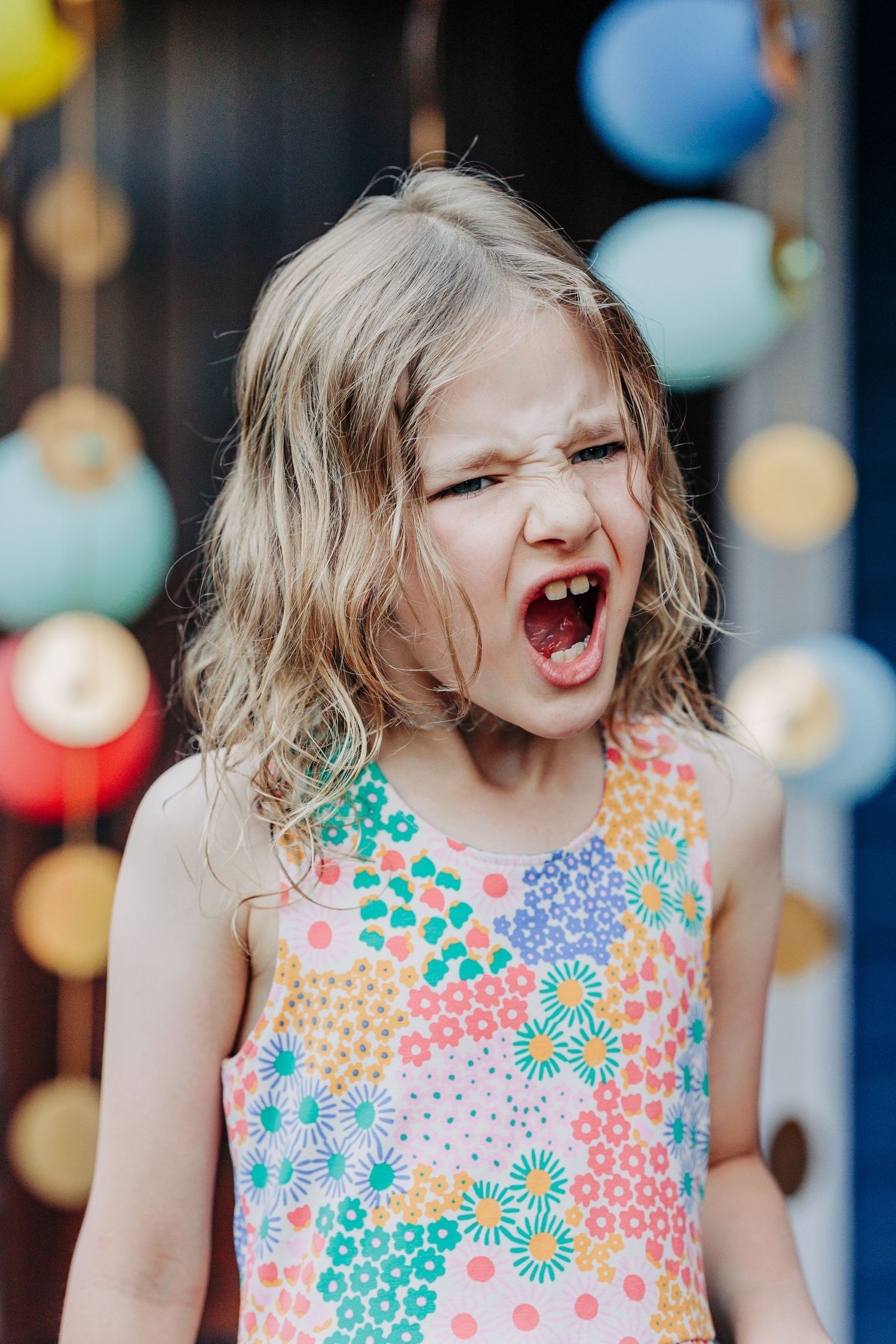 Expressive young girl in colorful floral dress roars playfully at outdoor birthday party with balloons.