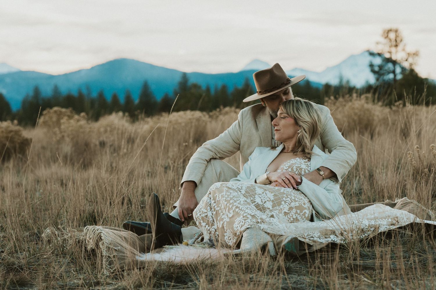 Romantic boho couple sitting together in a golden field with mountain views at dusk.