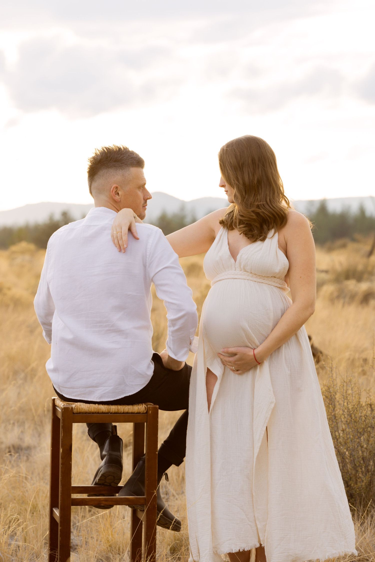 Pregnant woman in white dress resting hand on partner's shoulder during outdoor maternity shoot.