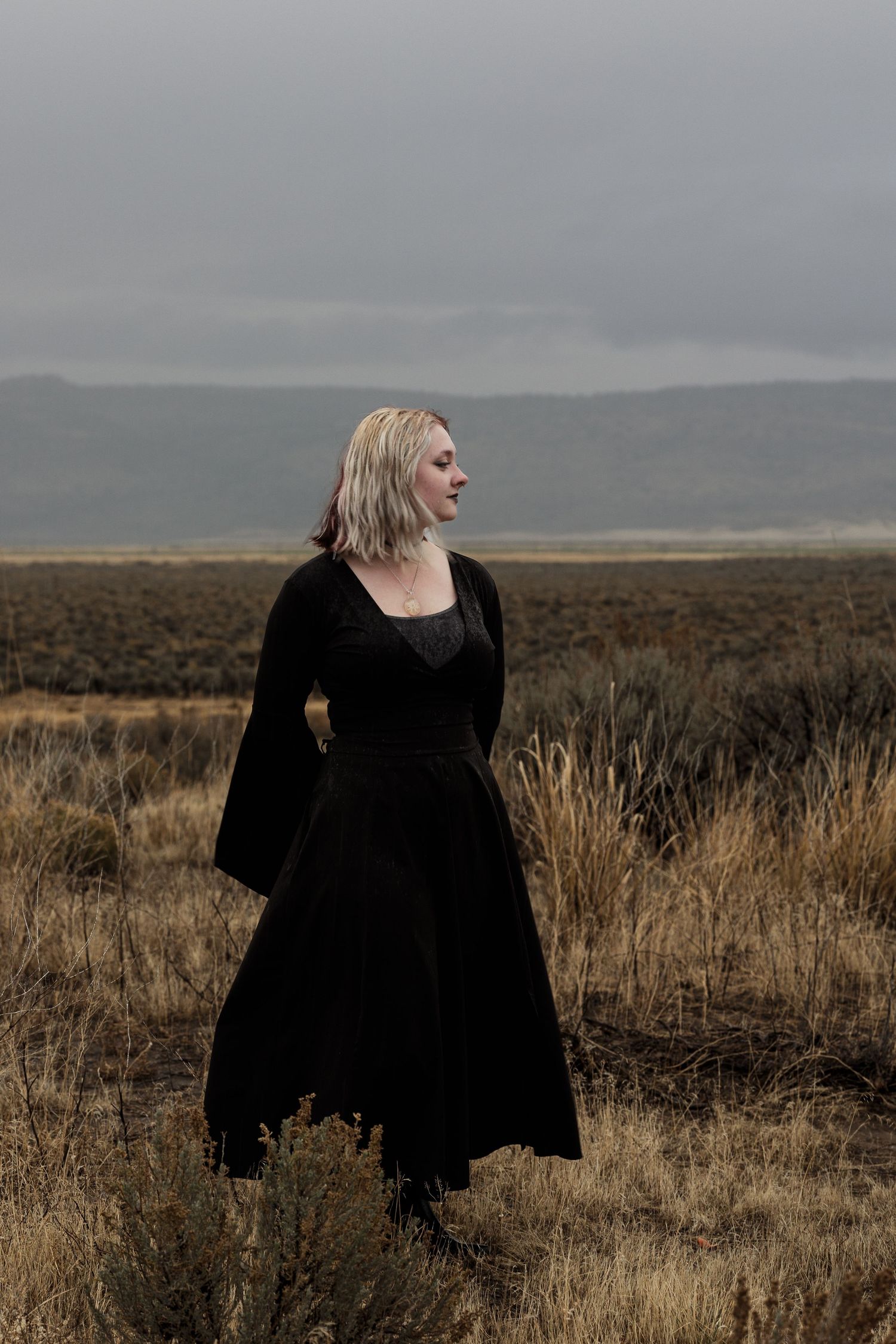 Woman in elegant black velvet dress standing alone in a dry desert field under overcast skies.