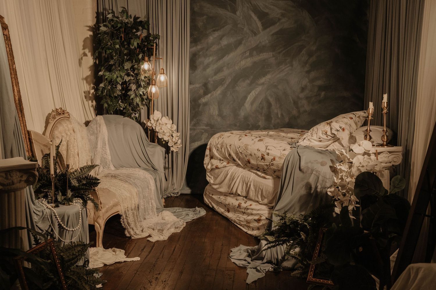Romantic bedroom setup with floral bedding, candles, lush greenery, and elegant vintage chair on dark hardwood floors.