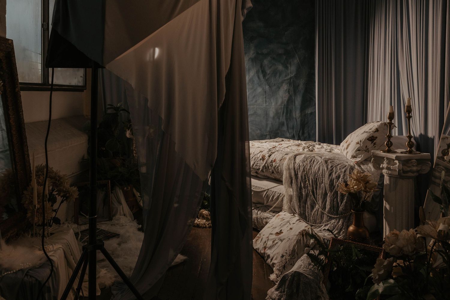 Moody bedroom photography studio setup with draped curtains, rumpled bed, and professional lighting equipment.
