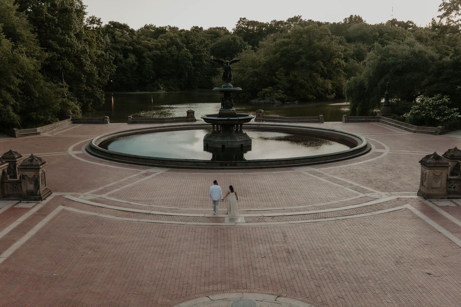 Couple holding hands at Bethesda Fountain in Central Park, NYC, surrounded by lush green trees.