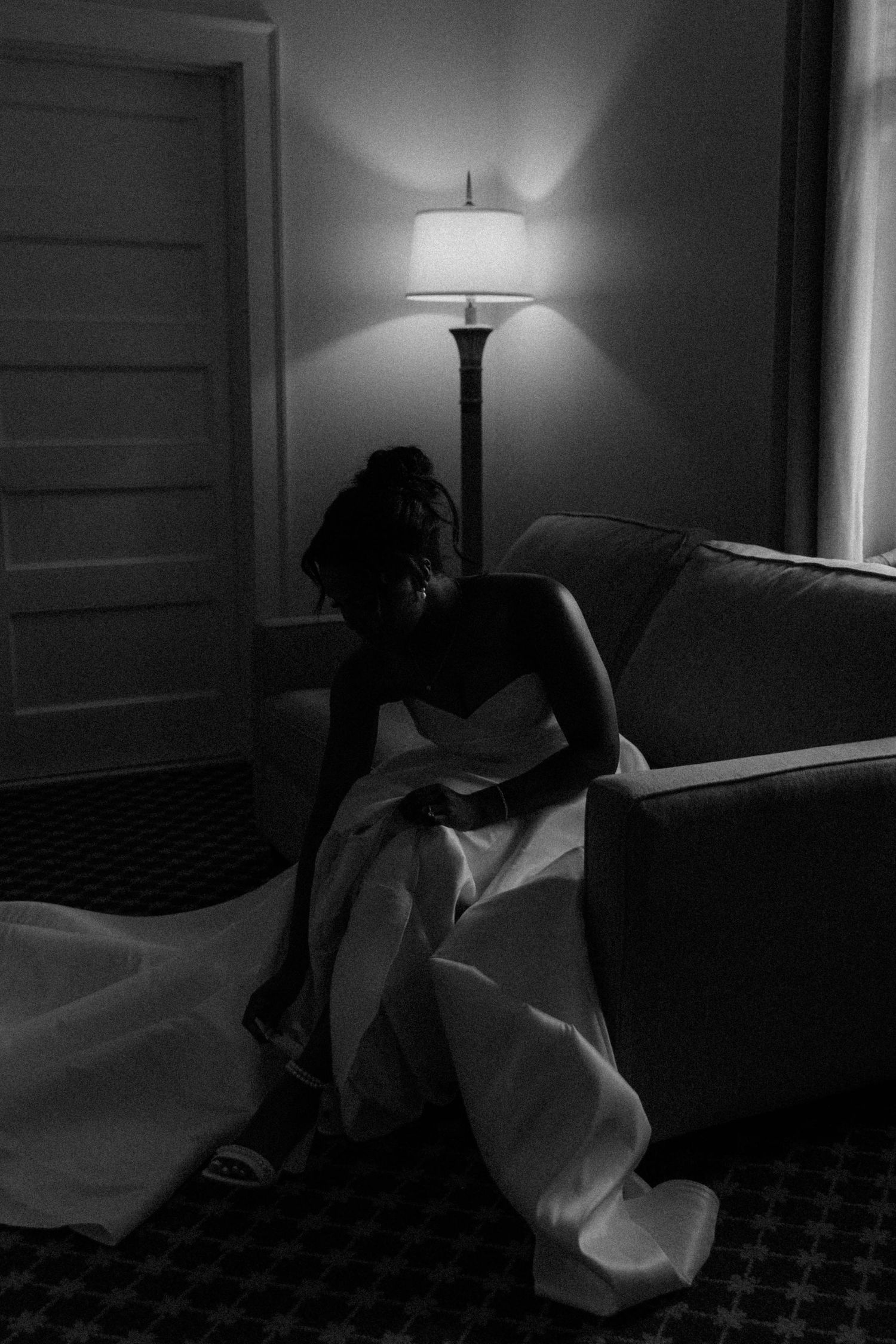 Black and white silhouette of bride in strapless gown sitting by a glowing floor lamp in hotel room.