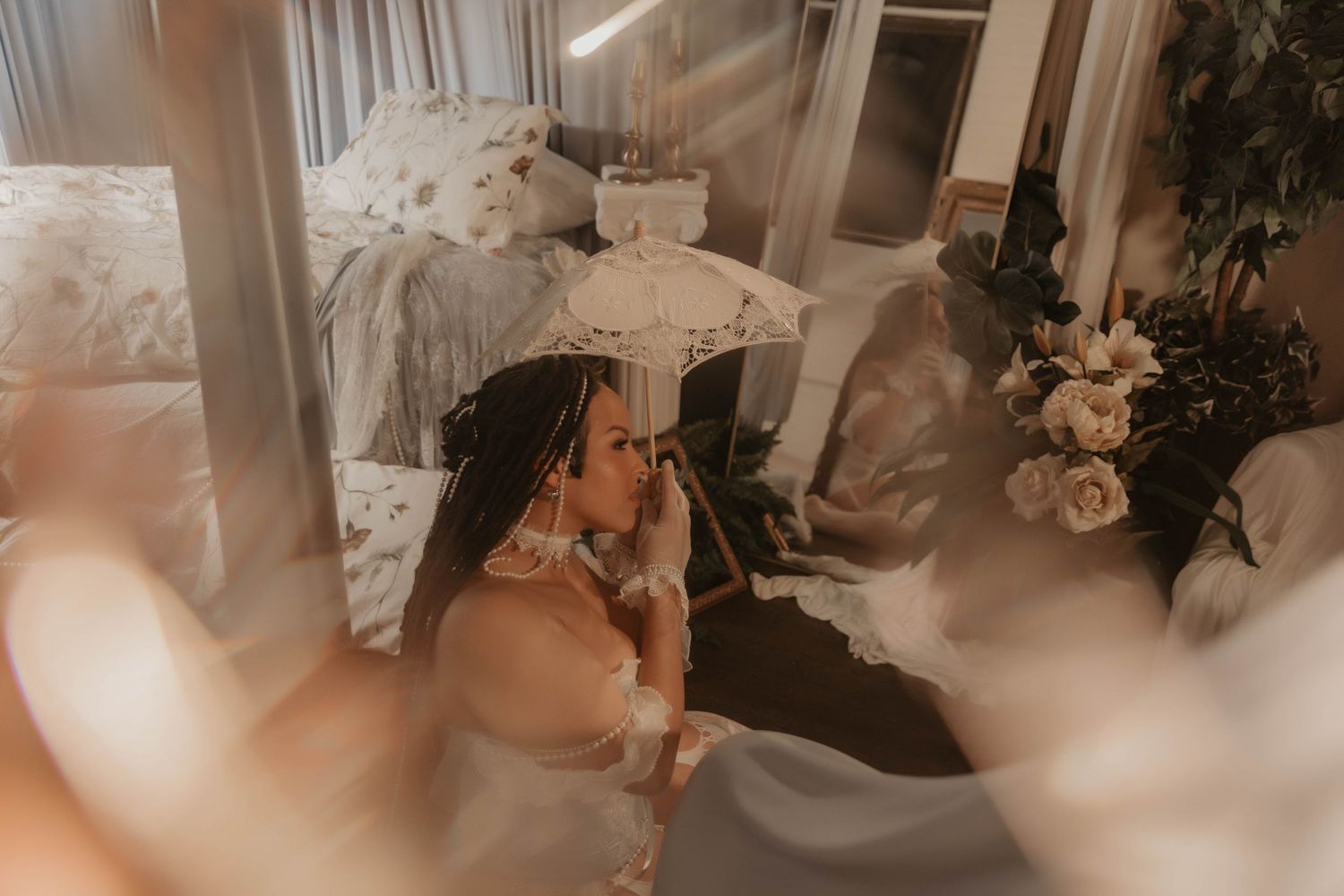 Bride in white gown holds lace parasol in dreamy, softly lit bridal boudoir with floral arrangements.