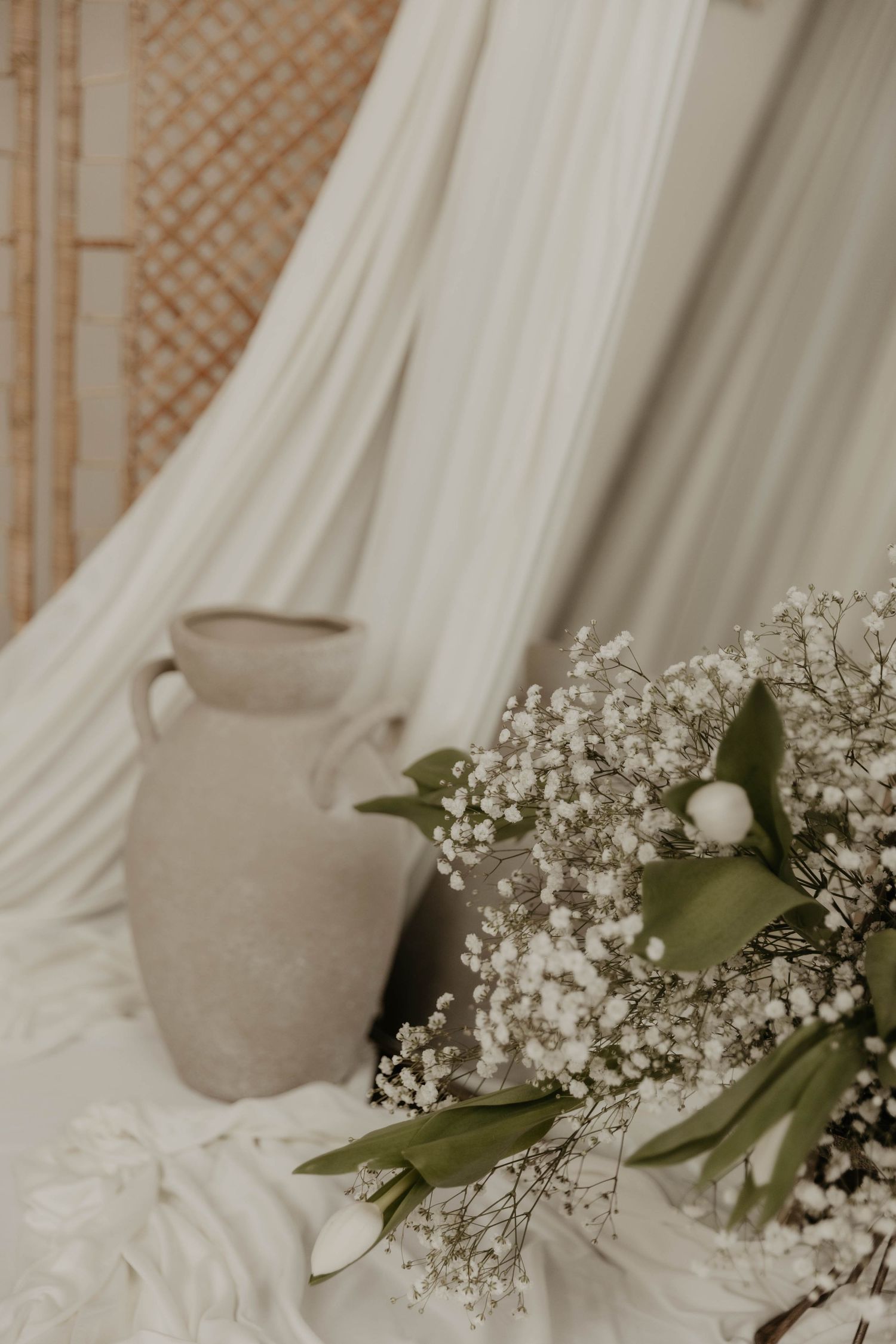 Boho wedding decor with white gypsophila bouquet, green leaves, and a rustic clay vase against draped white fabric.