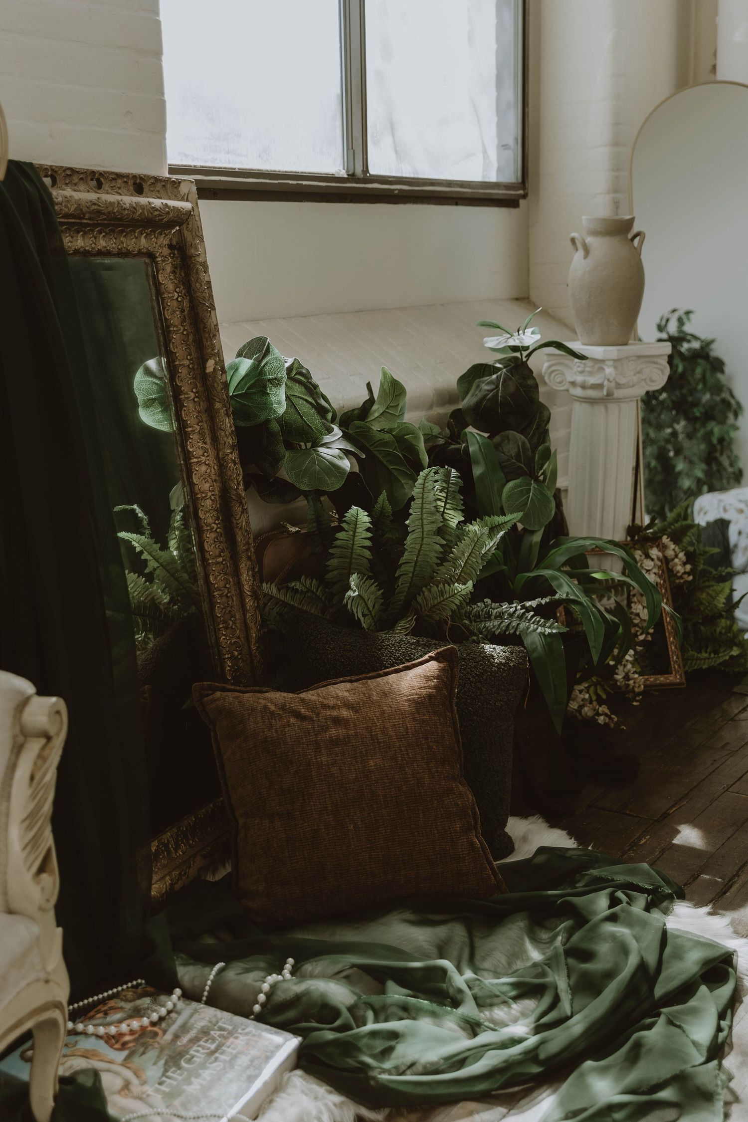 Boho interior corner with lush green plants, ornate gold mirror, brown velvet pillow, and green fabric on the floor.
