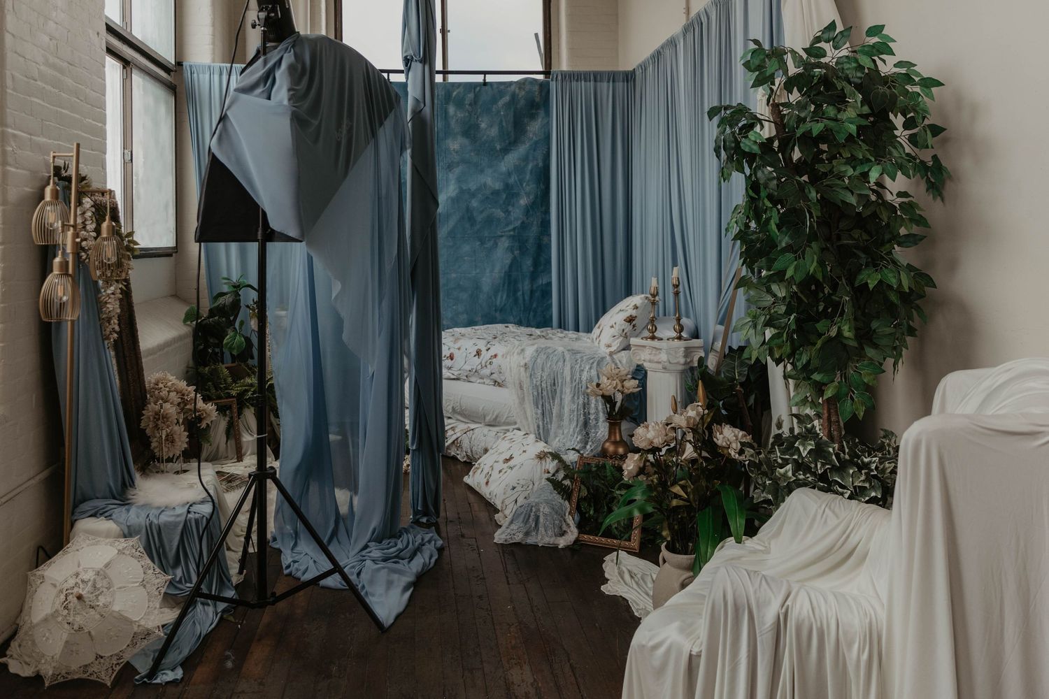 A styled photography studio with blue fabric draping, a bed with floral decor, plants, and soft white coverings.