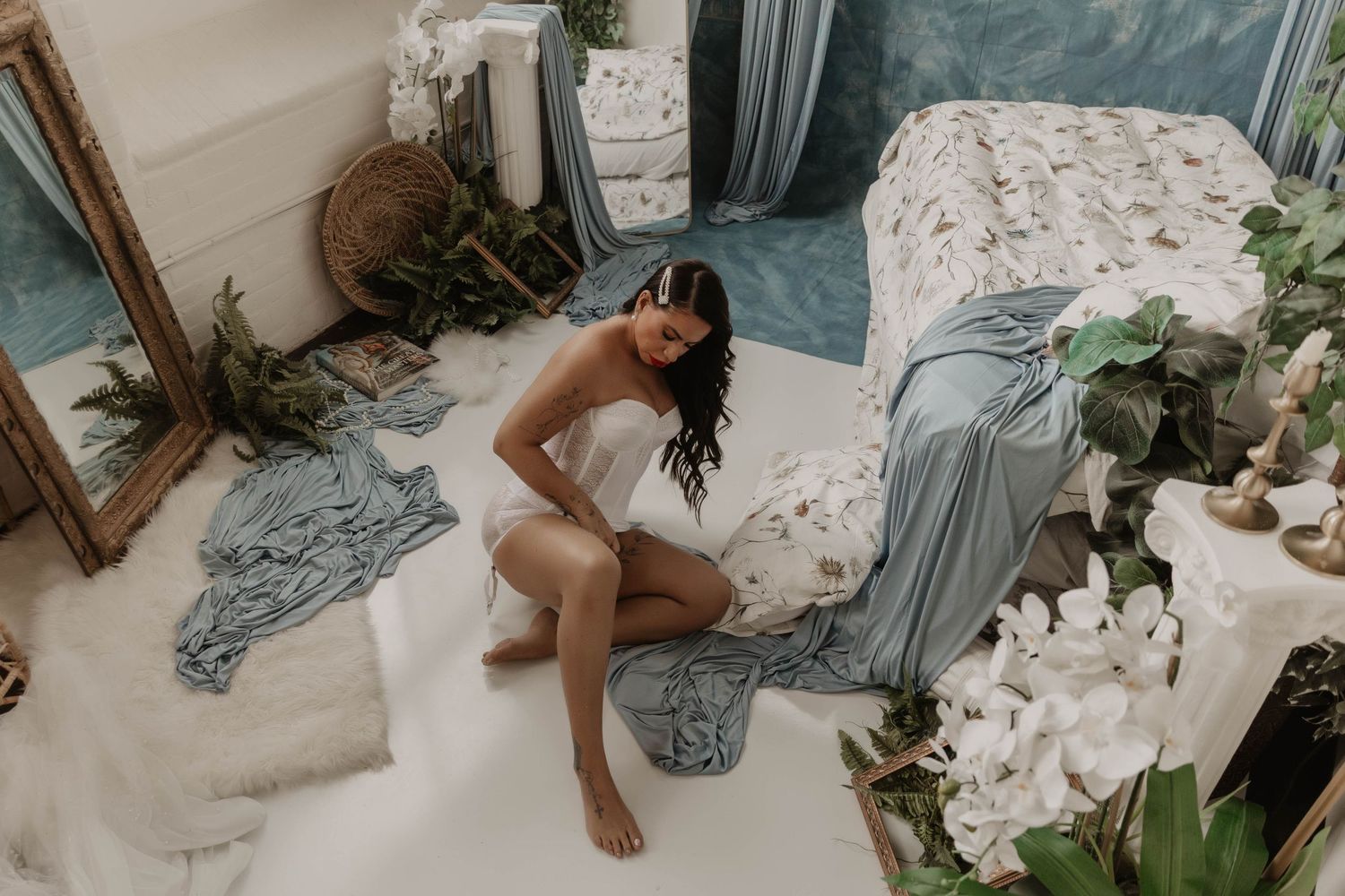 A woman in white lingerie sits gracefully on the floor of a dreamy blue and white bedroom studio setting.