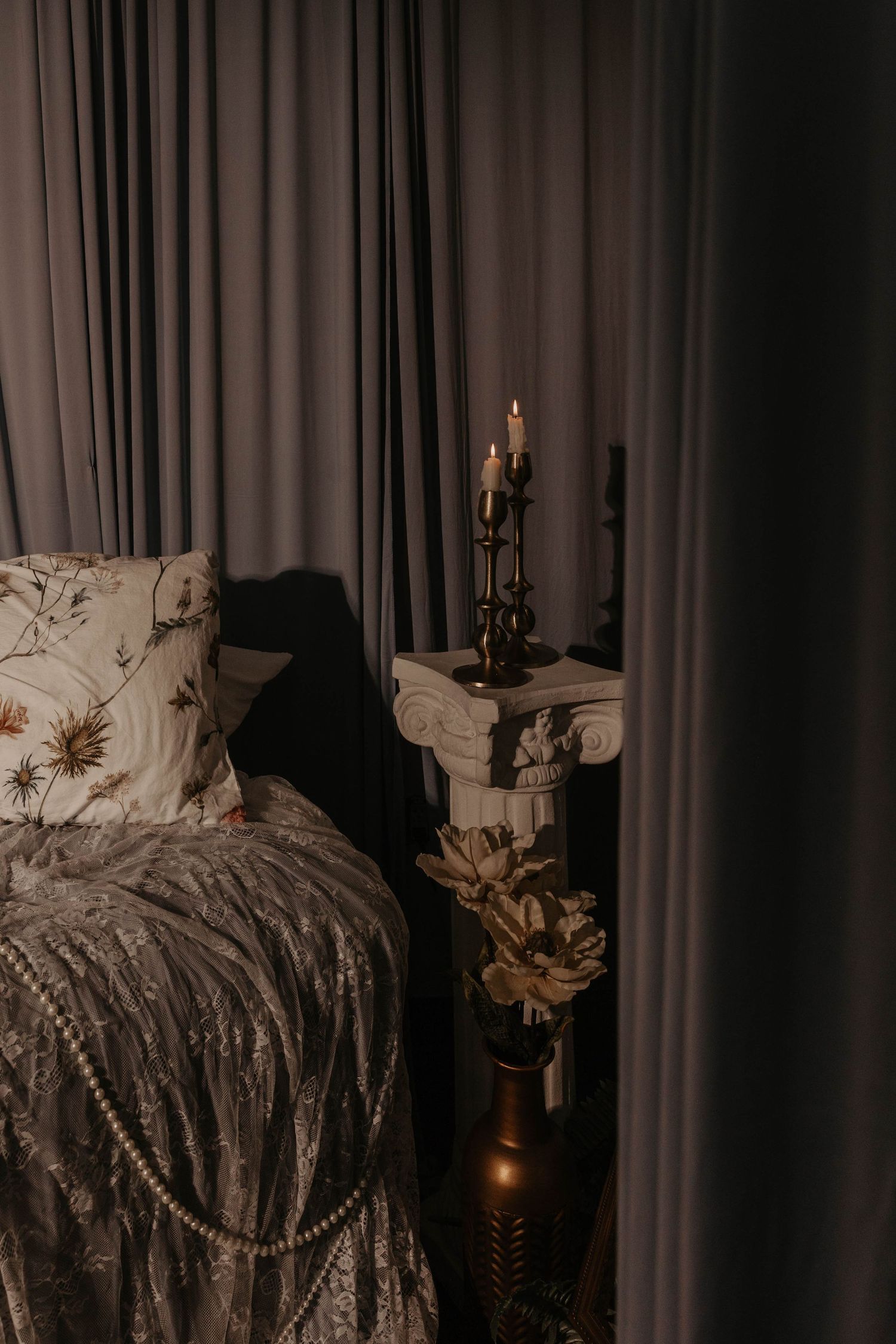 Dark romantic bedroom with lit candles on ornate pedestal, floral pillows, gray velvet bedding, and gold vase beside draped curtains.