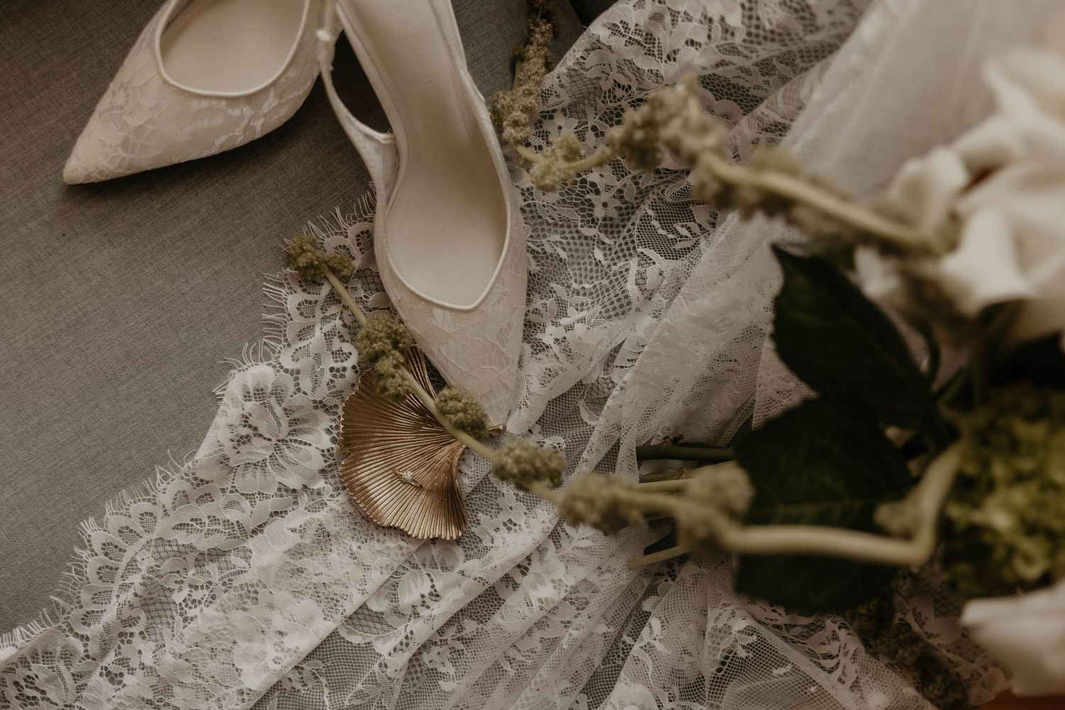 Elegant bridal details featuring lace wedding dress, ivory heels, and delicate floral accents arranged on soft fabric.