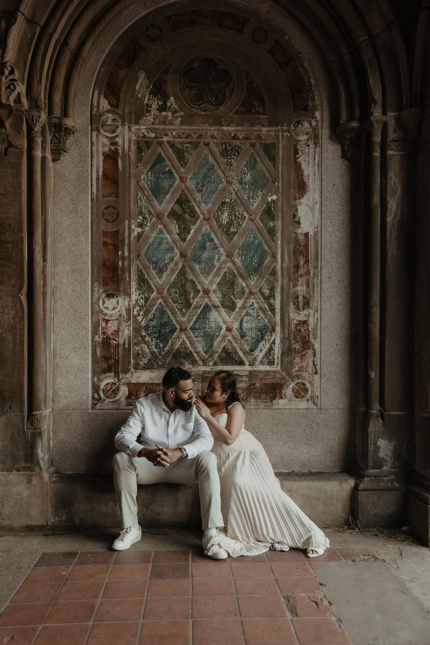 Couple in white wedding attire sitting together under ornate arch with colorful mosaic backdrop at Bethesda Terrace.