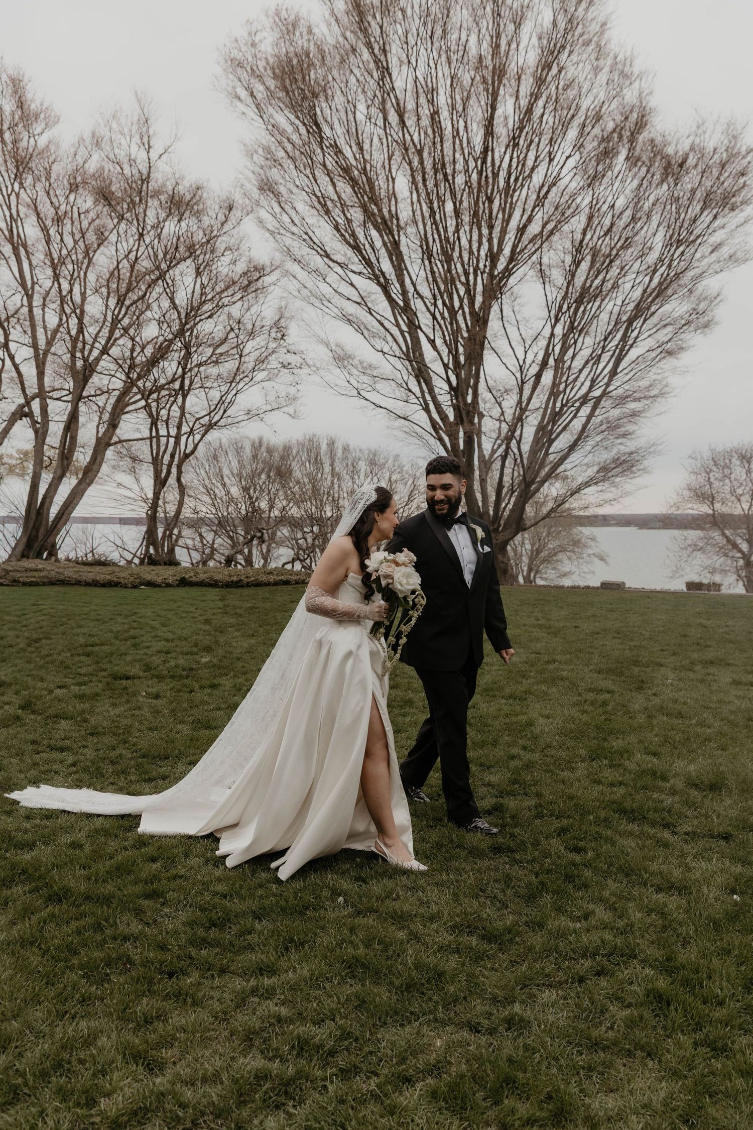 Bride and groom walk across green lawn with bare winter trees and river in background at wedding.

top rated RI wedding photographer