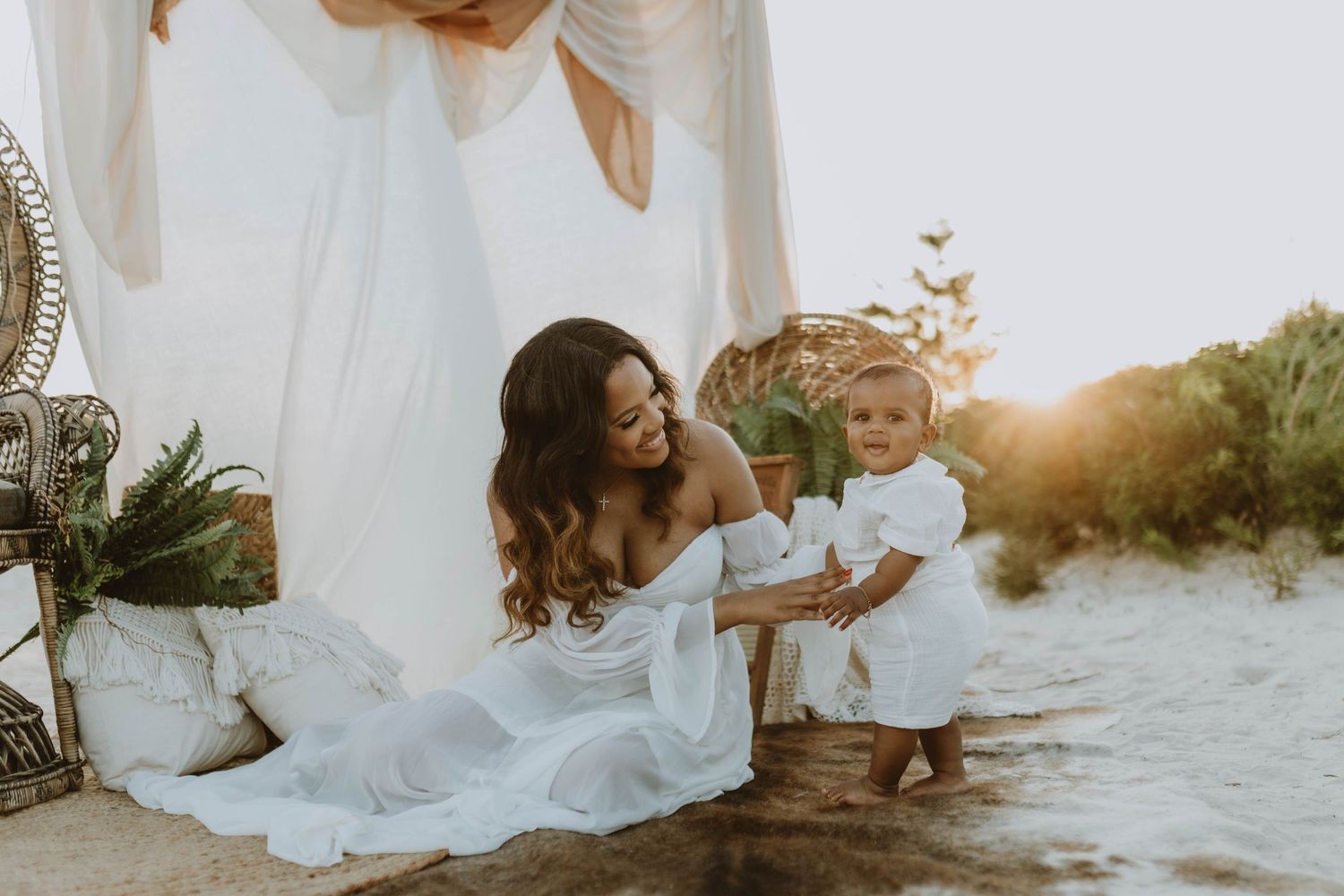 A mother in a white dress sits on a beach at sunset smiling at her baby in a white outfit standing beside her.