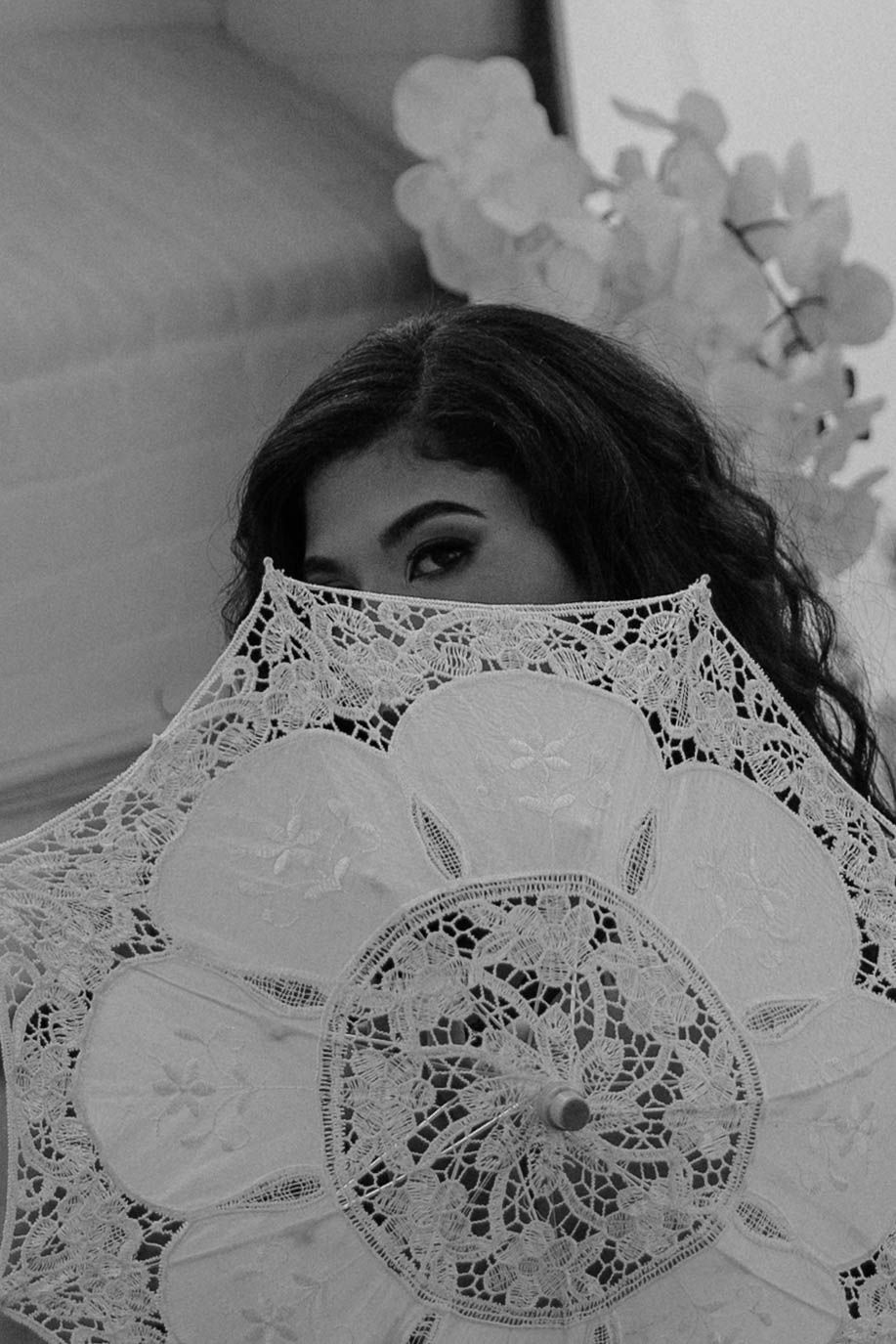 Black and white photo of a woman peeking over an ornate lace parasol umbrella with floral decorations in background.