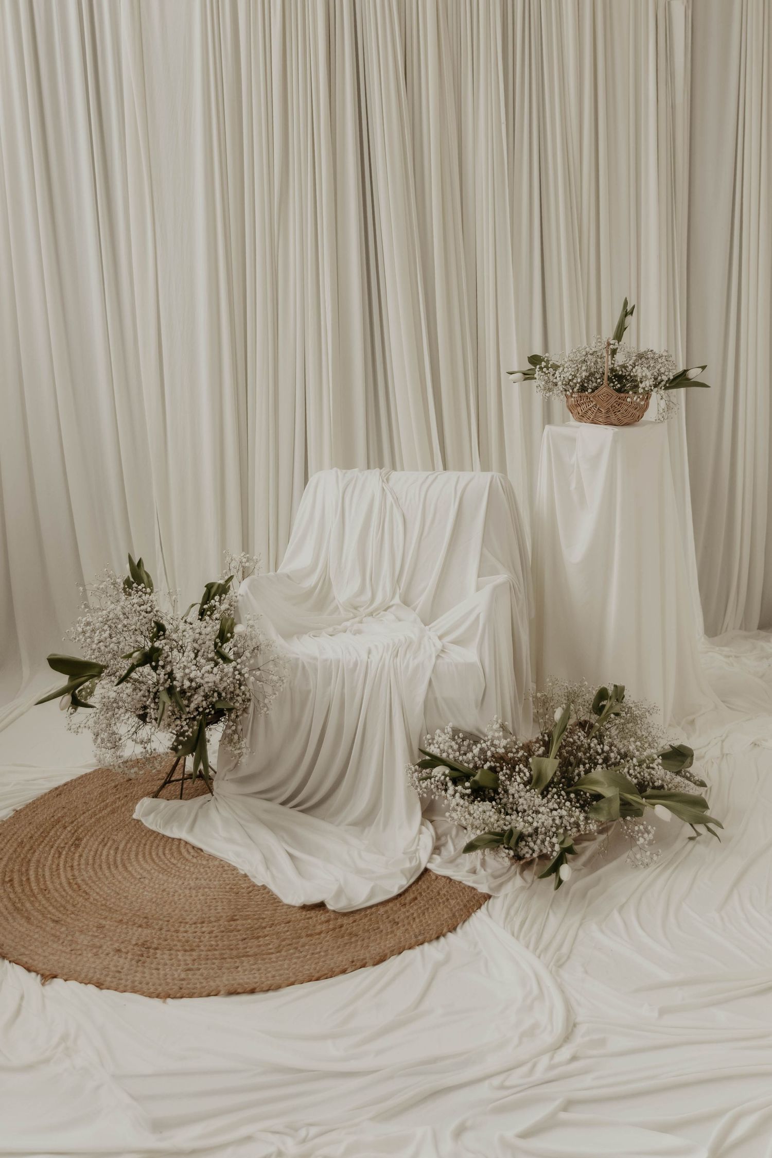 Elegant white draped chair with floral arrangements on a jute rug against white curtain backdrop.