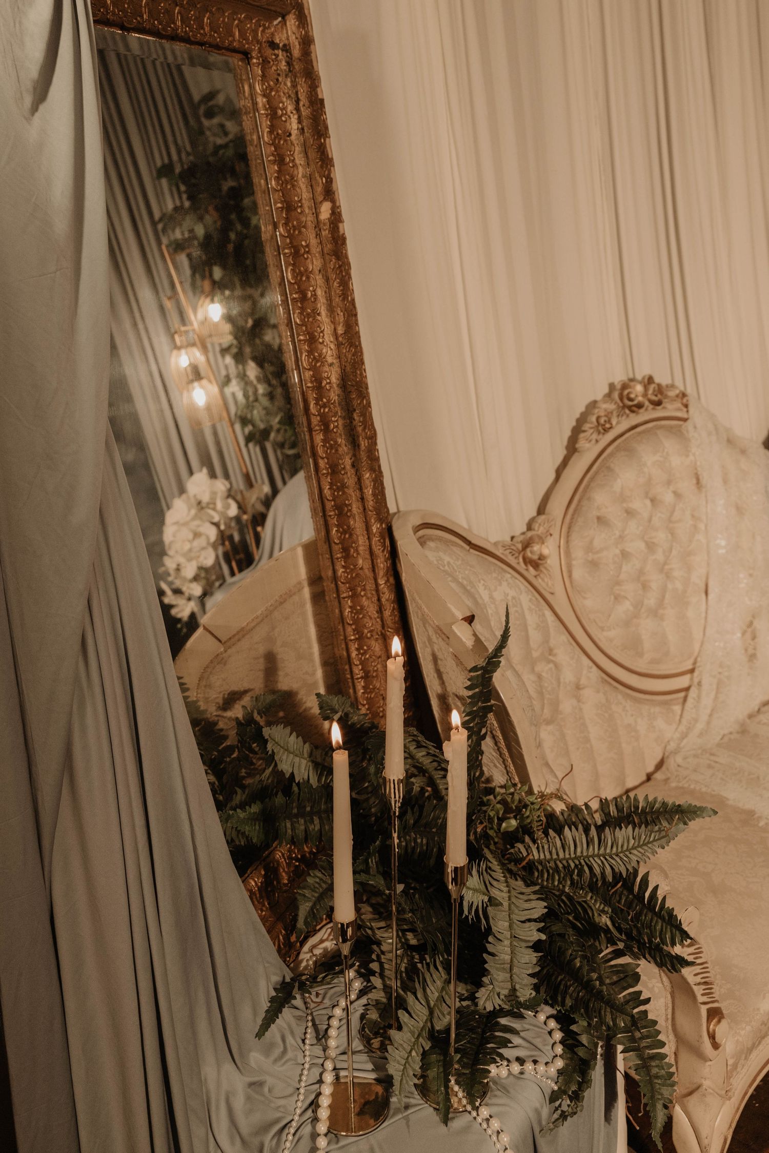 Elegant gold-framed mirror with lit candles, fern greenery, and ornate white chair in a romantic wedding decor setting.