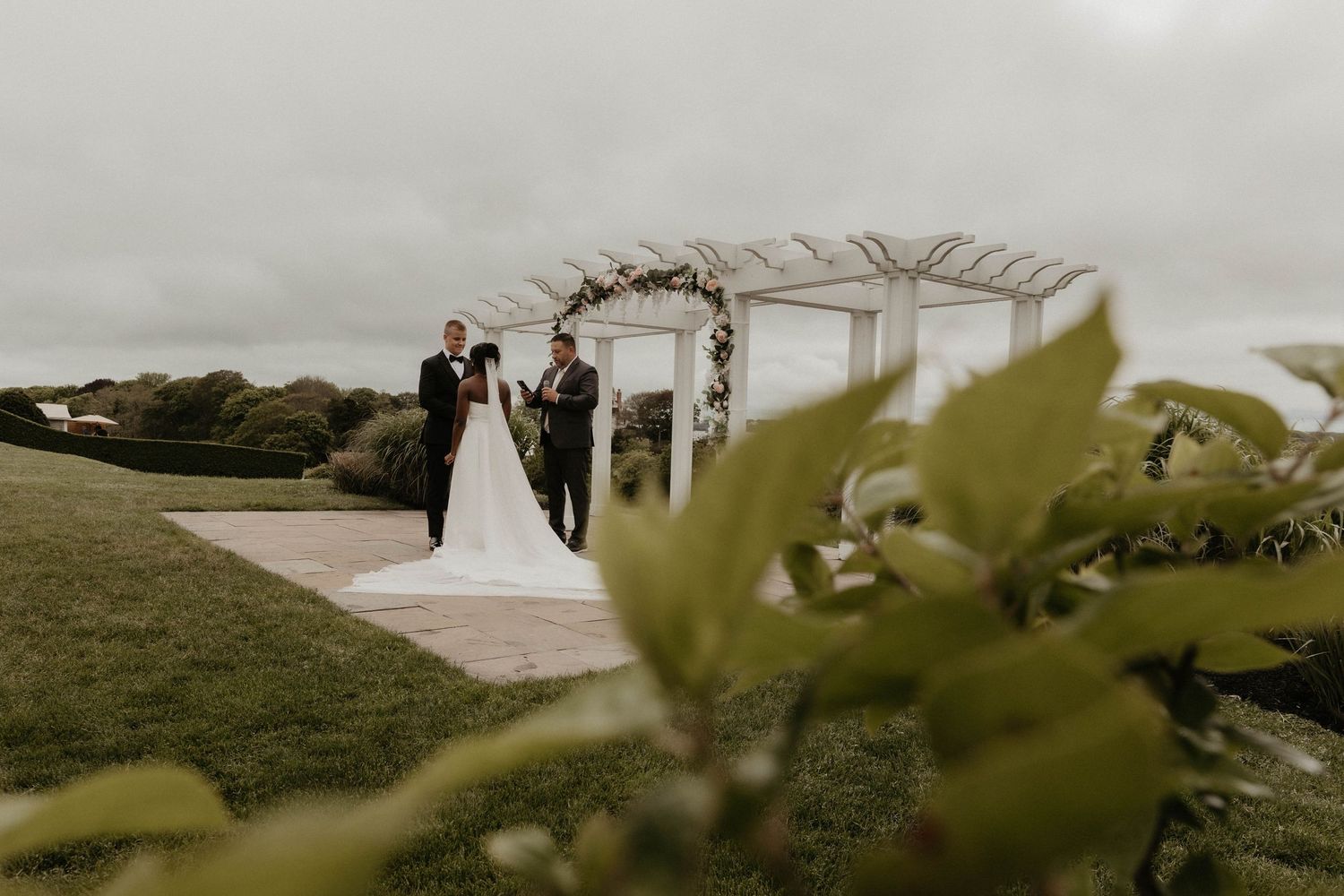Bride and groom exchanging vows at a floral arch pergola ceremony on an overcast outdoor wedding day.