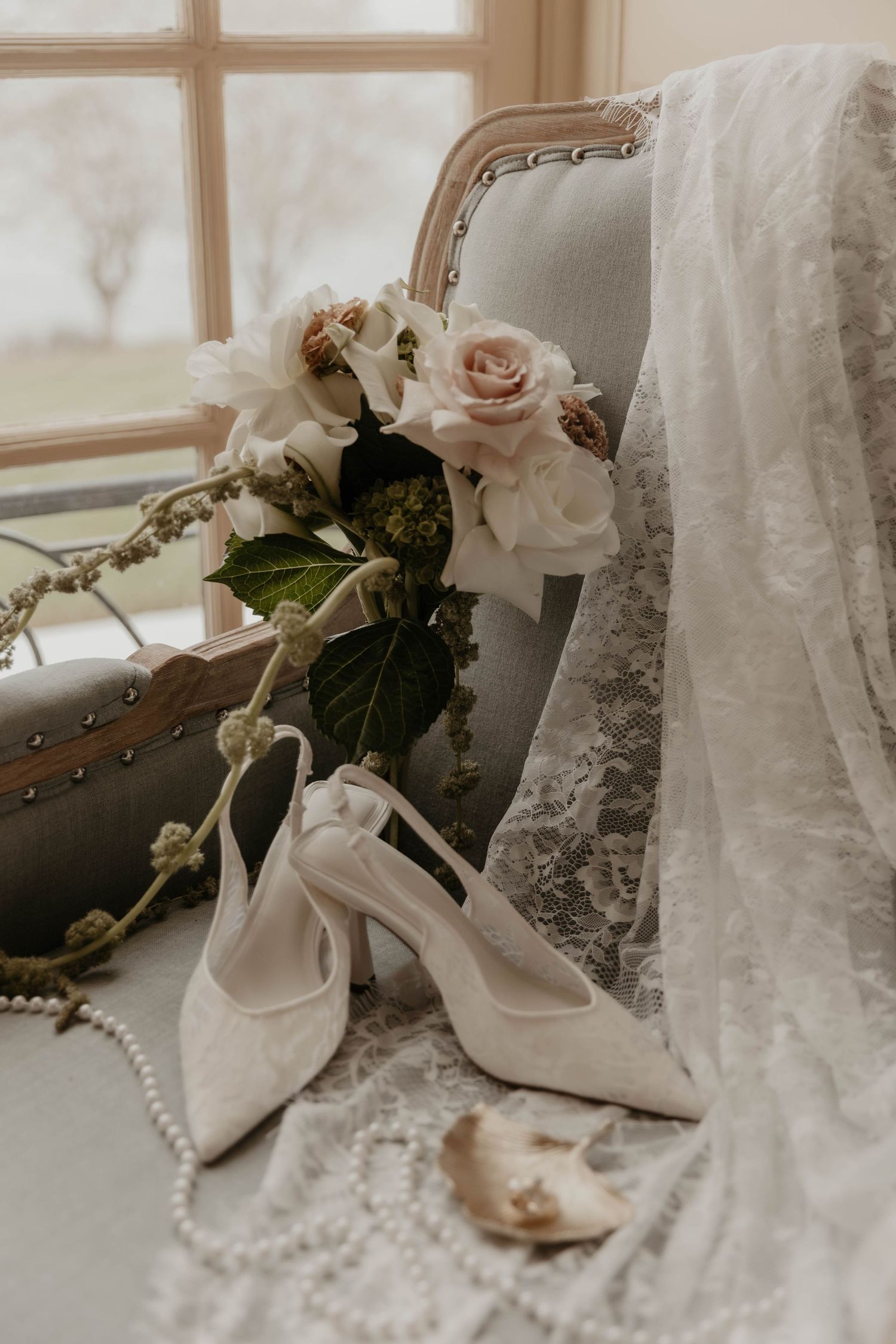 White bridal heels with pearl details beside pink roses and lace veil on vintage chair by window.

top rated RI wedding photographer