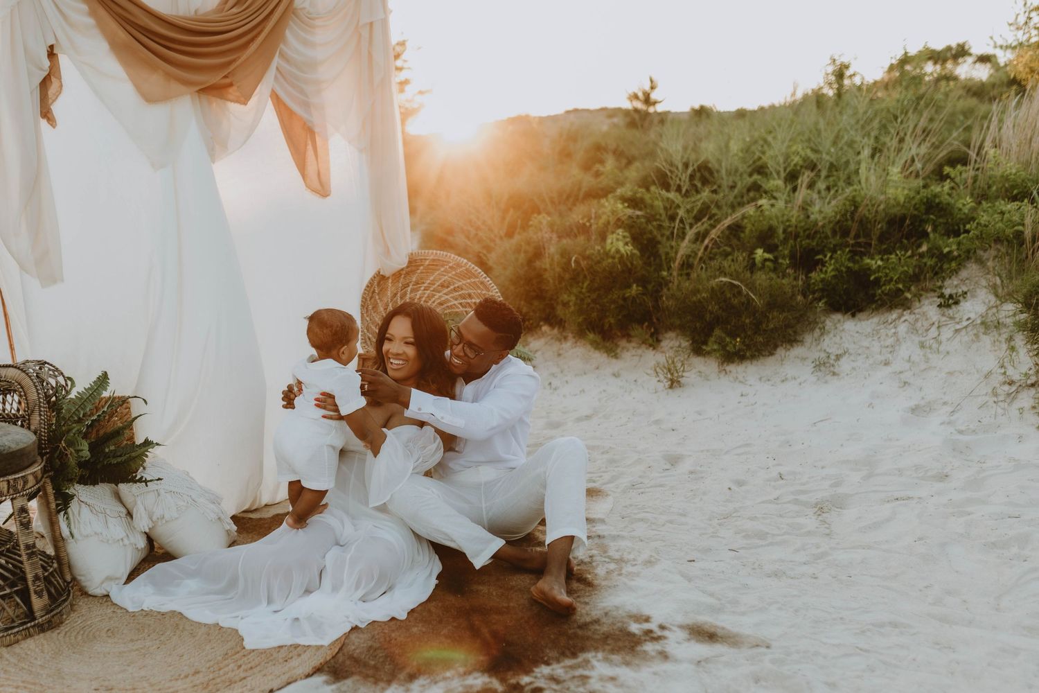 A Black family in white outfits sits on sandy beach at golden hour, parents lovingly holding their baby near dune grass.