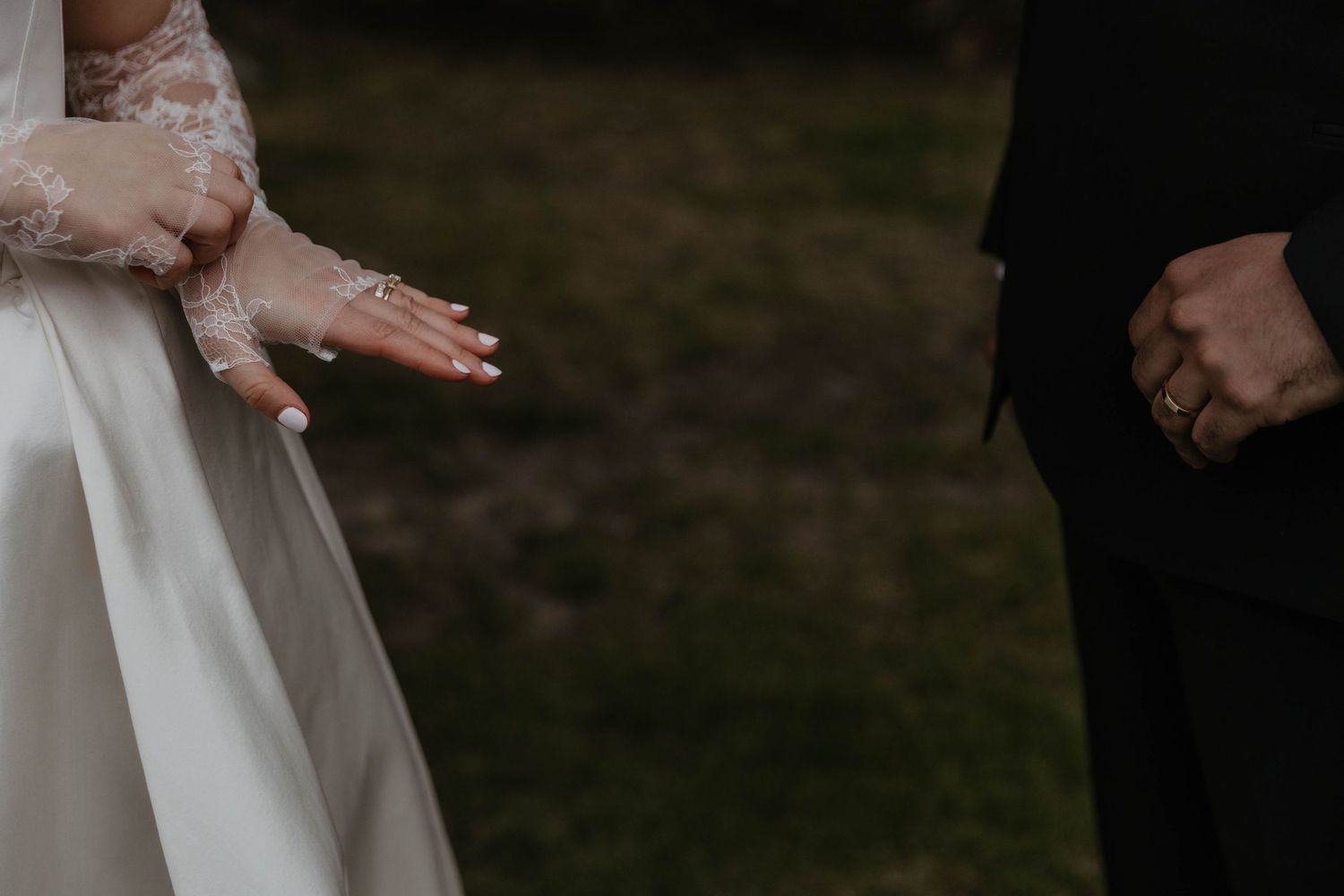 Close-up of bride in lace wedding gown extending hand toward groom in black suit outdoors.
