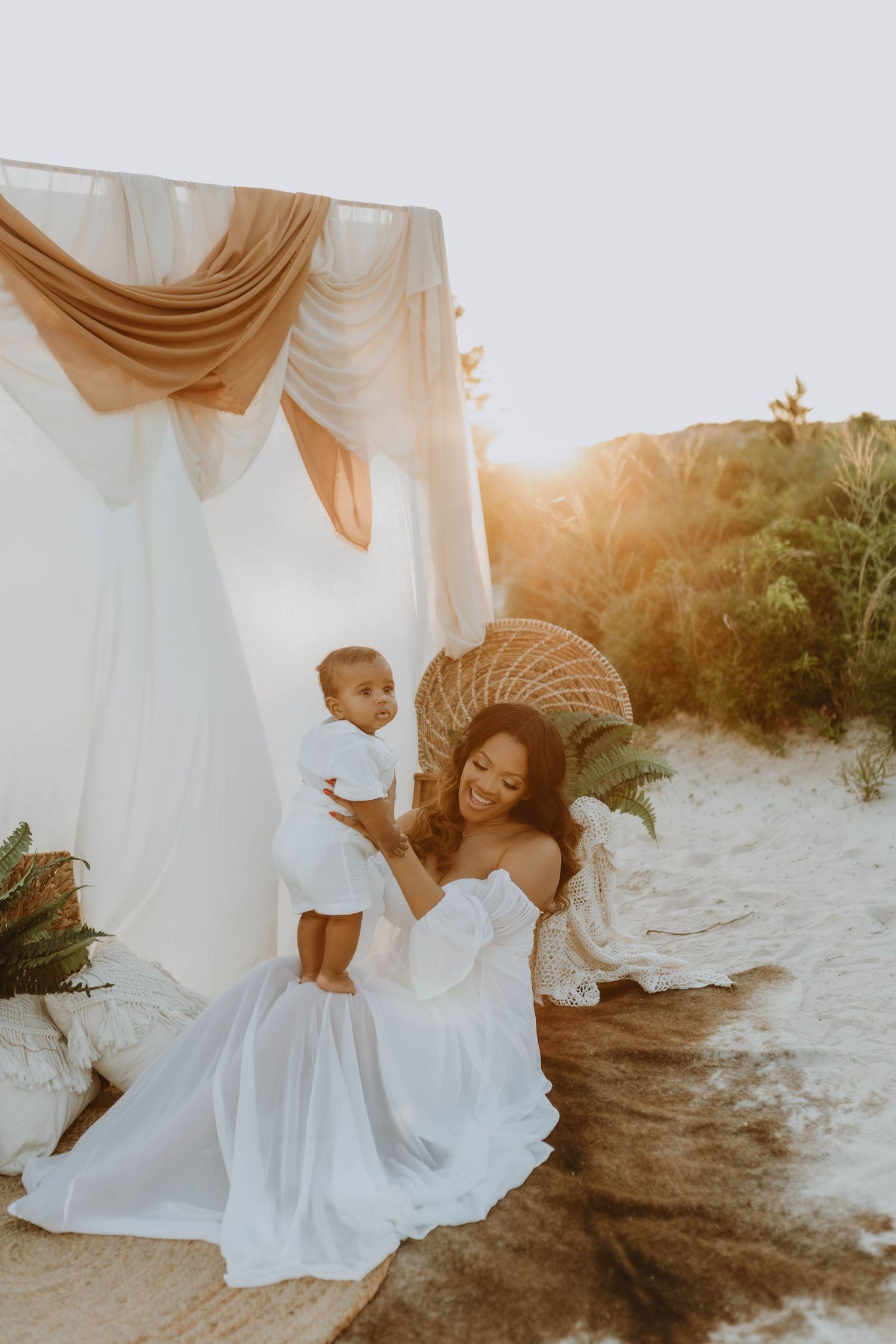Mother in white gown holds baby at golden hour boho beach setup with draped fabric canopy and wicker decor.