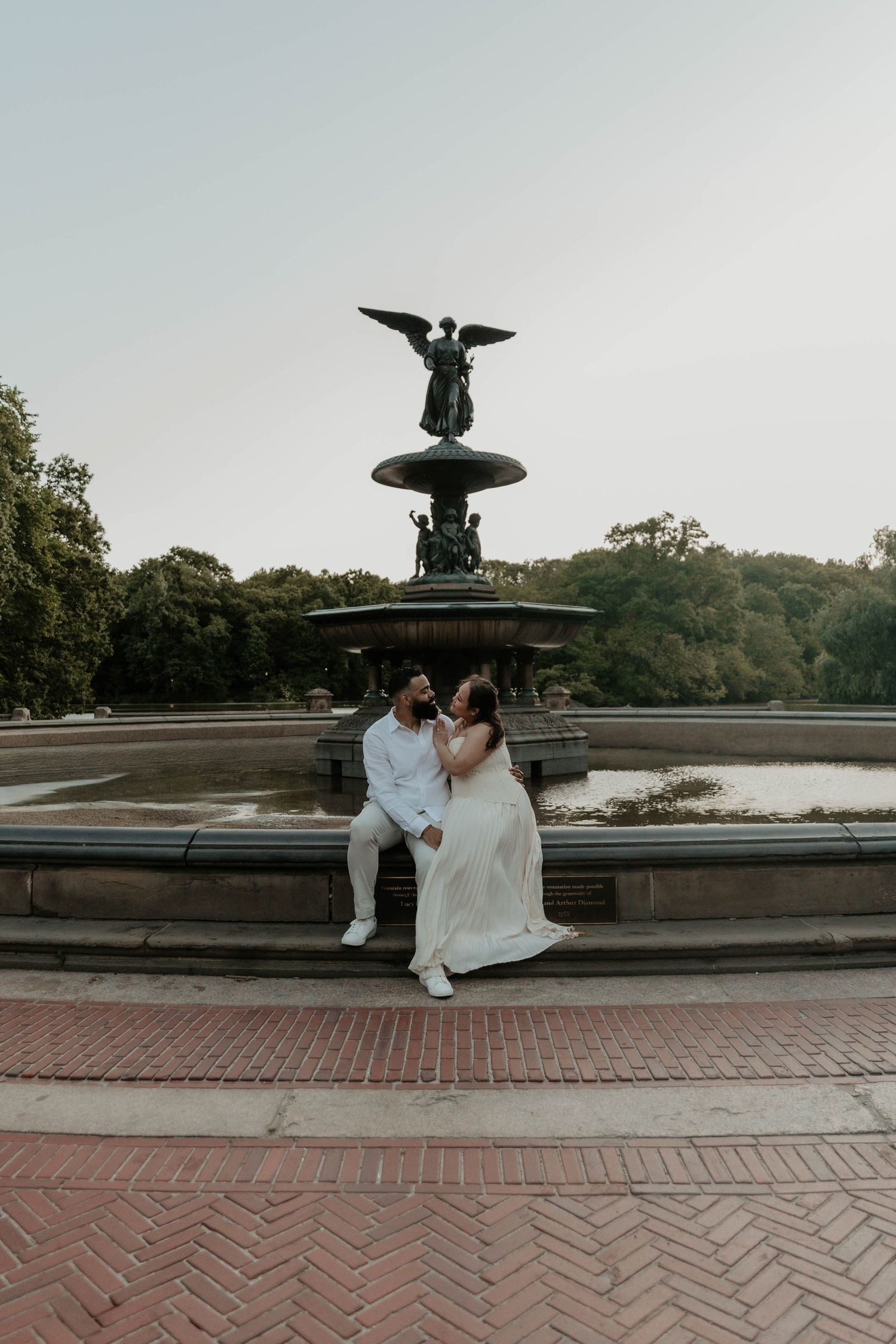 Couple in wedding attire sitting together at Bethesda Fountain in Central Park, NYC, with angel statue backdrop.