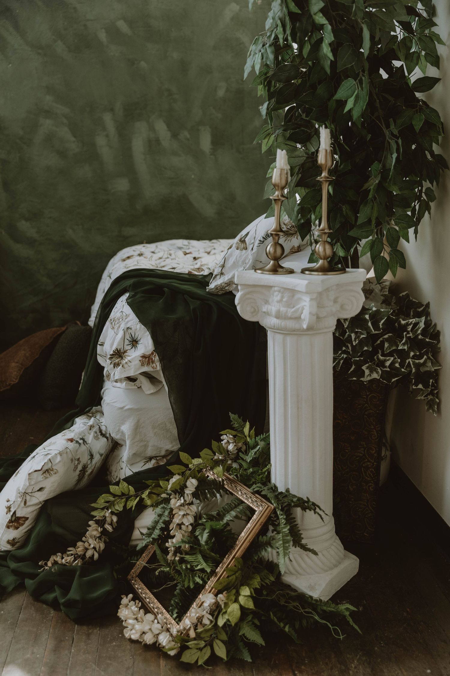 Elegant wedding decor with white column, gold candlesticks, greenery, floral pillows, and ornate gold frame on wooden floor.