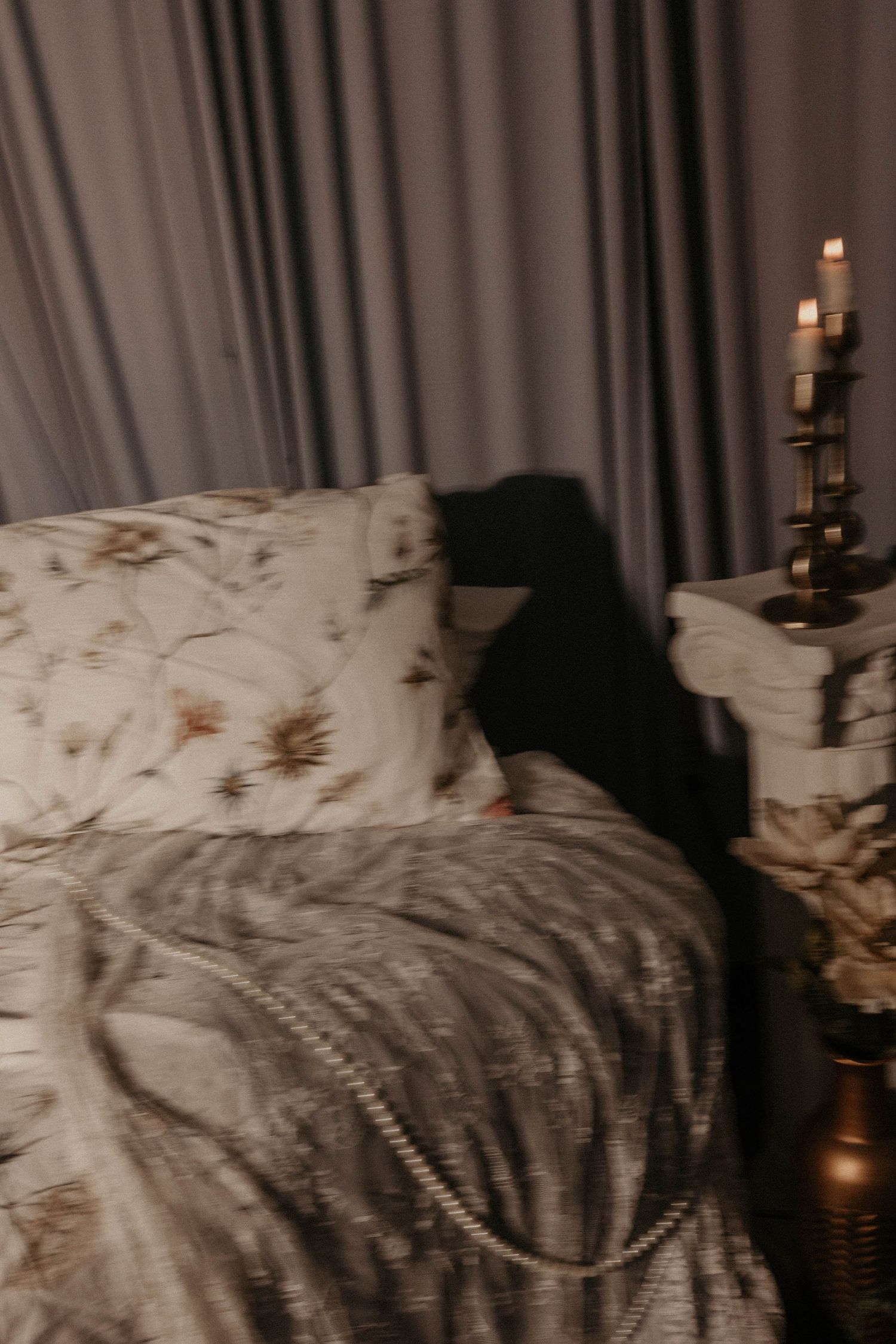 Cozy dark bedroom with floral pillows, gray velvet blanket, pearl necklace, and lit candles on a vintage nightstand.