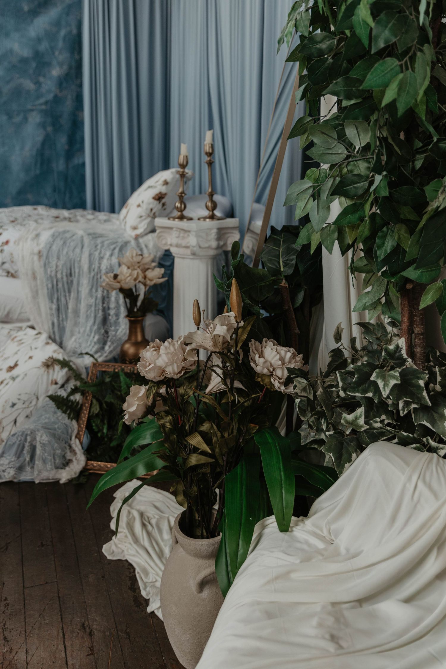 Elegant room with blue curtains, white flowers, green plants, candles on pedestal, and draped white fabric on wood floor.
