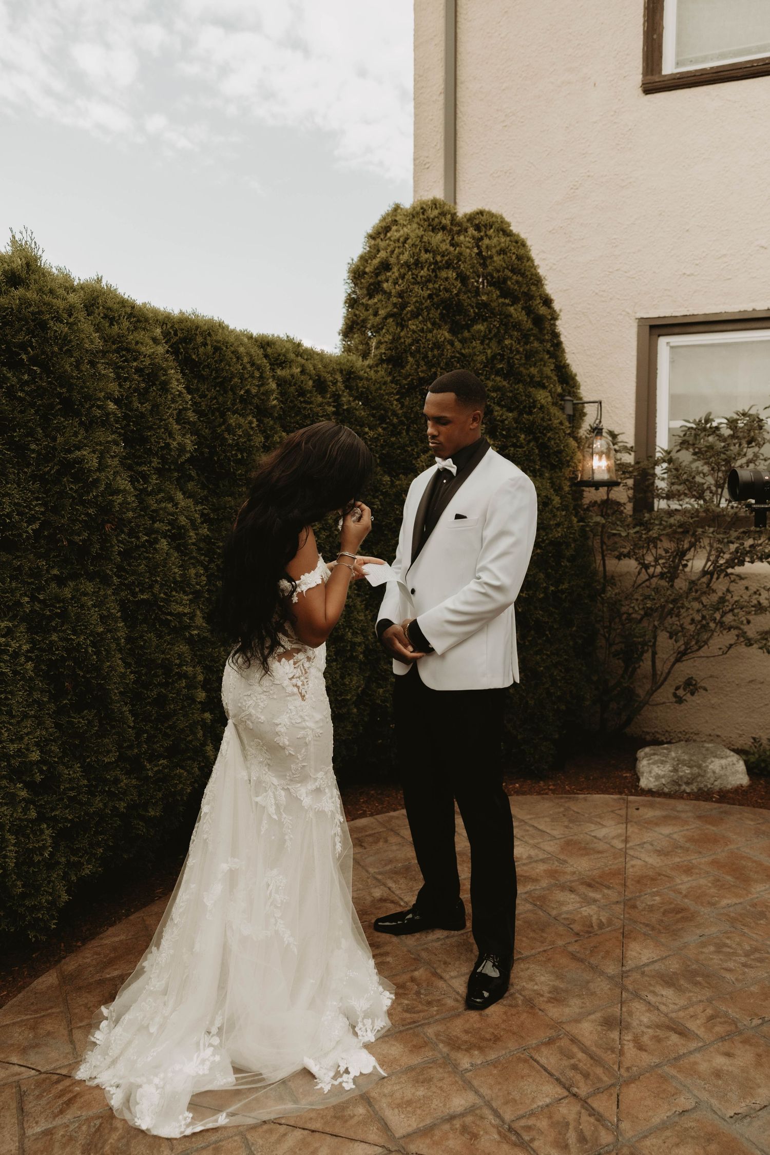 Bride in lace mermaid gown and groom in white tuxedo sharing an intimate moment in outdoor courtyard.
