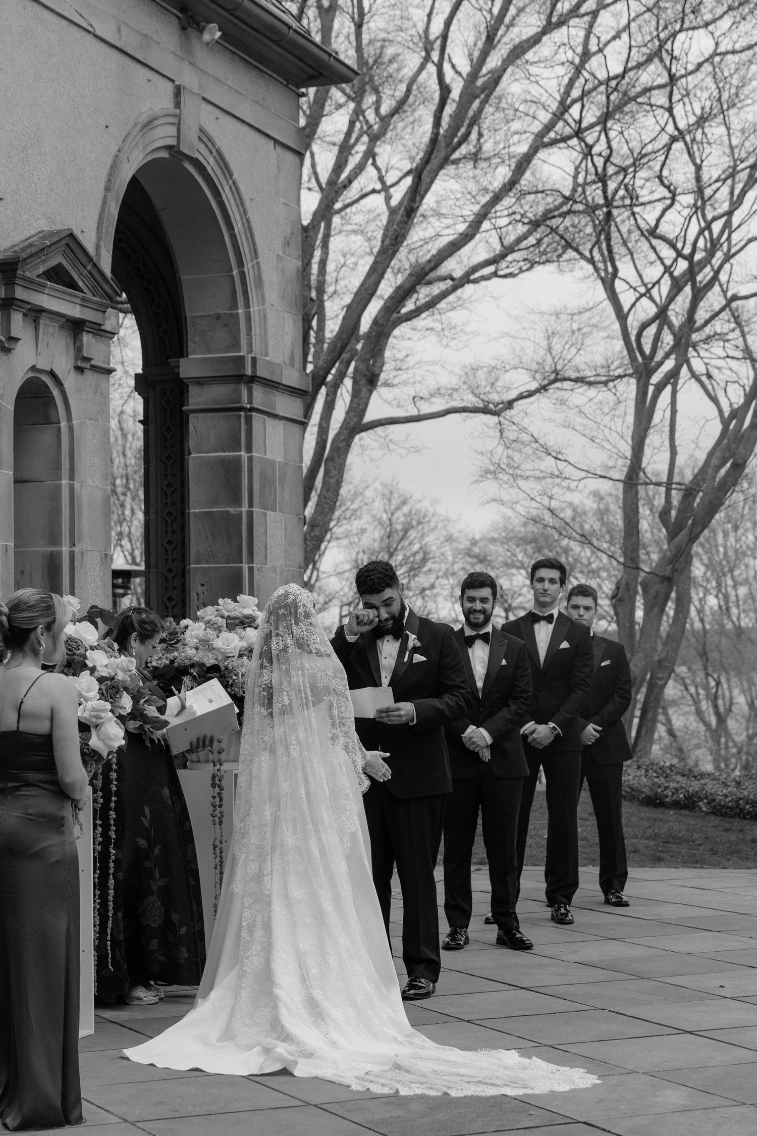 Black and white outdoor wedding ceremony with groom reading vows before groomsmen at stone venue.

top rated RI wedding photographer