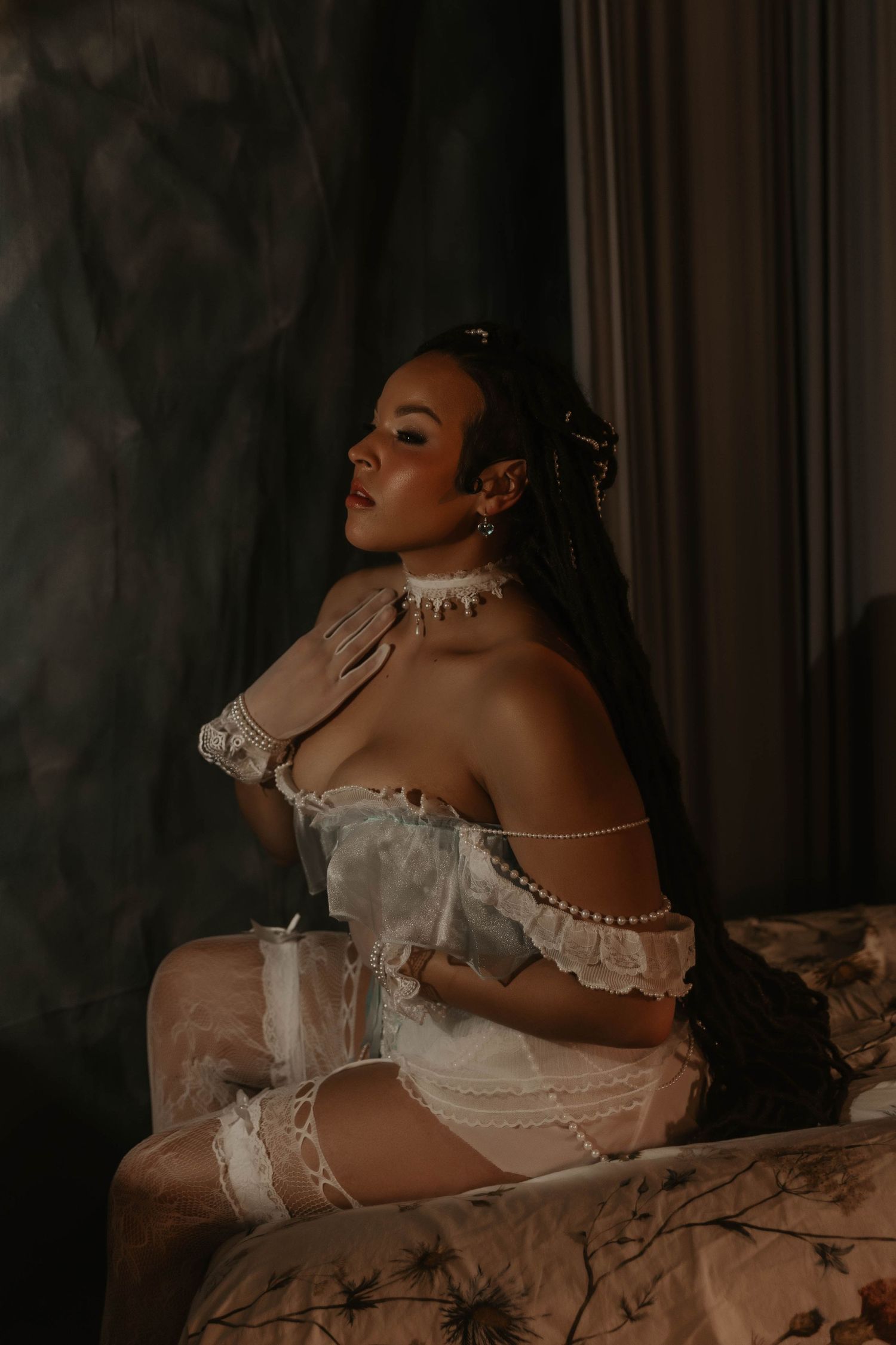 Woman in white lace lingerie and pearl accessories poses elegantly on a bed in moody, dramatic lighting.
