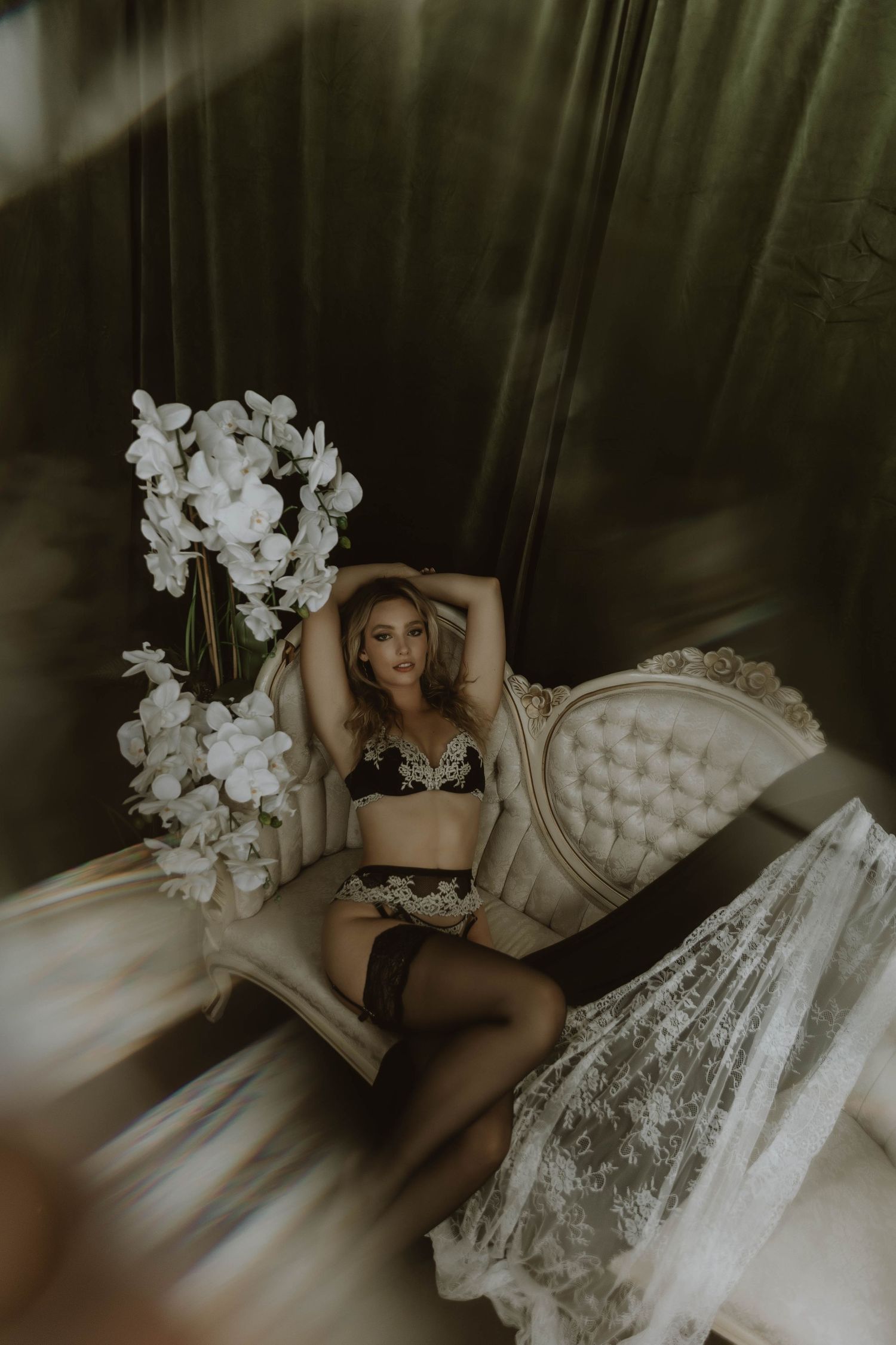 Blonde woman in black embroidered lingerie and stockings posing on vintage chair with white orchids.