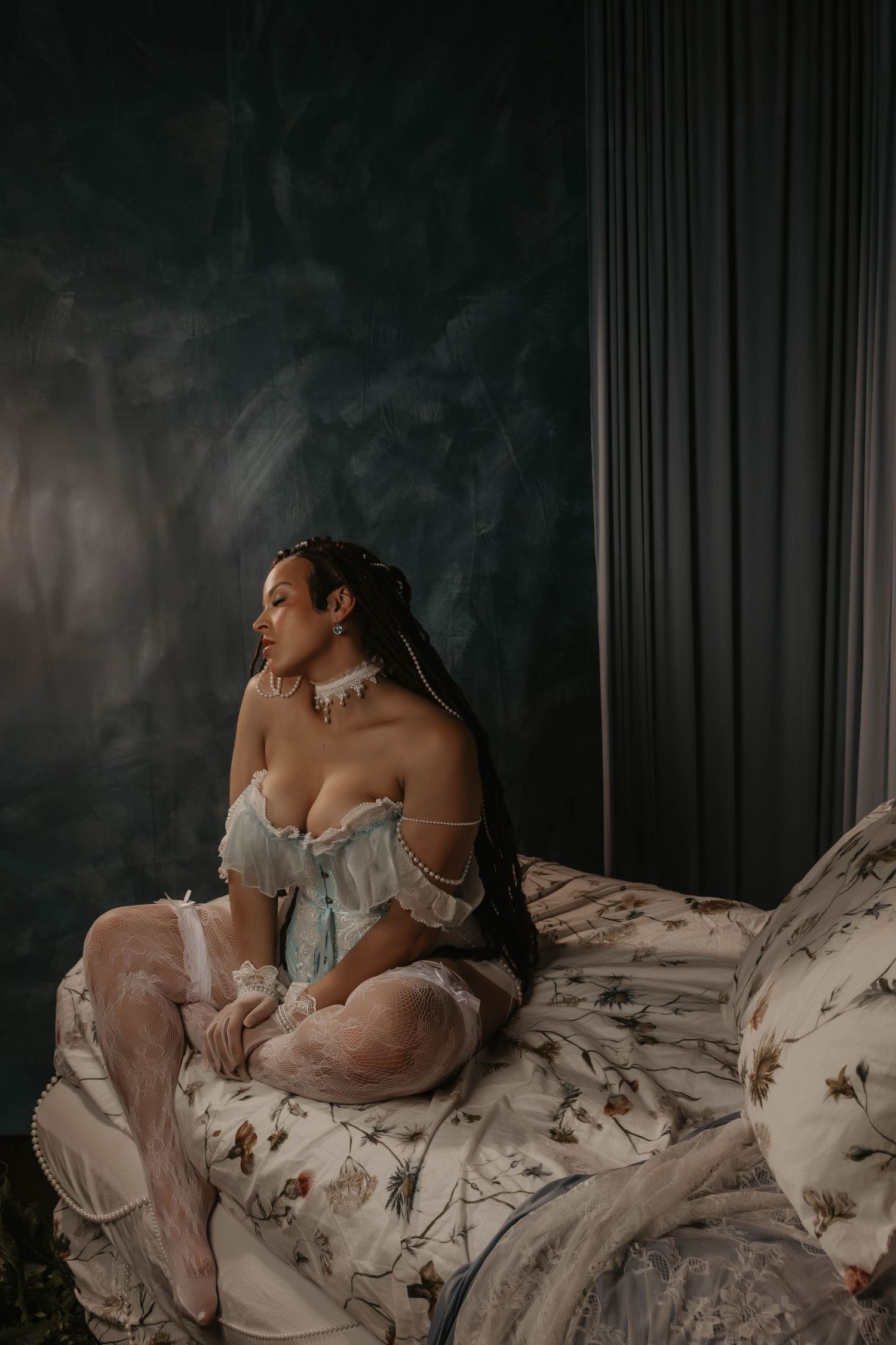 Woman in elegant off-shoulder corset sits gracefully on floral bedding against a moody dark teal backdrop.
